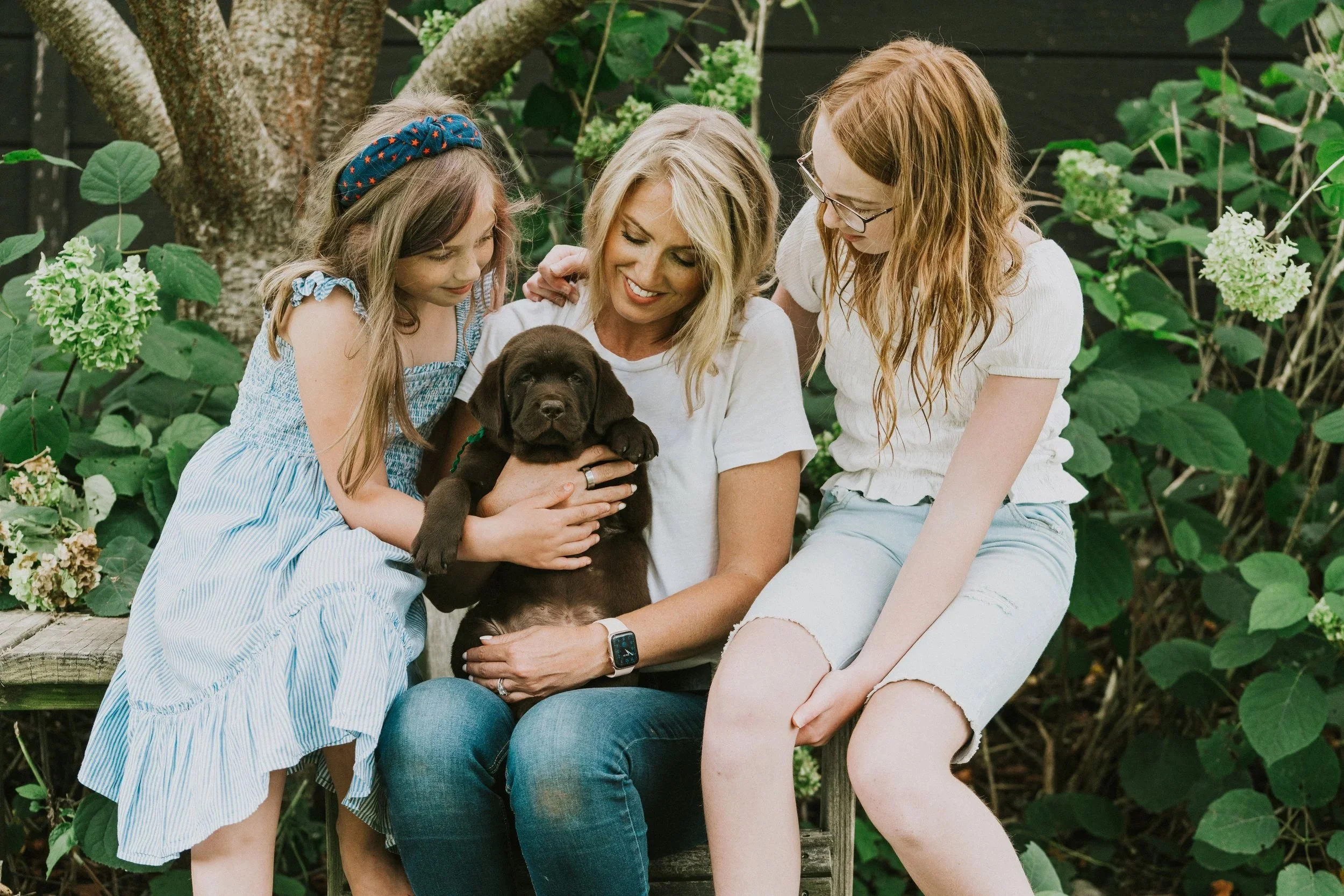 CopperRidge family sitting outdoors, smiling and holding a chocolate Labrador puppy. They are surrounded by green plants and a tree in the background.