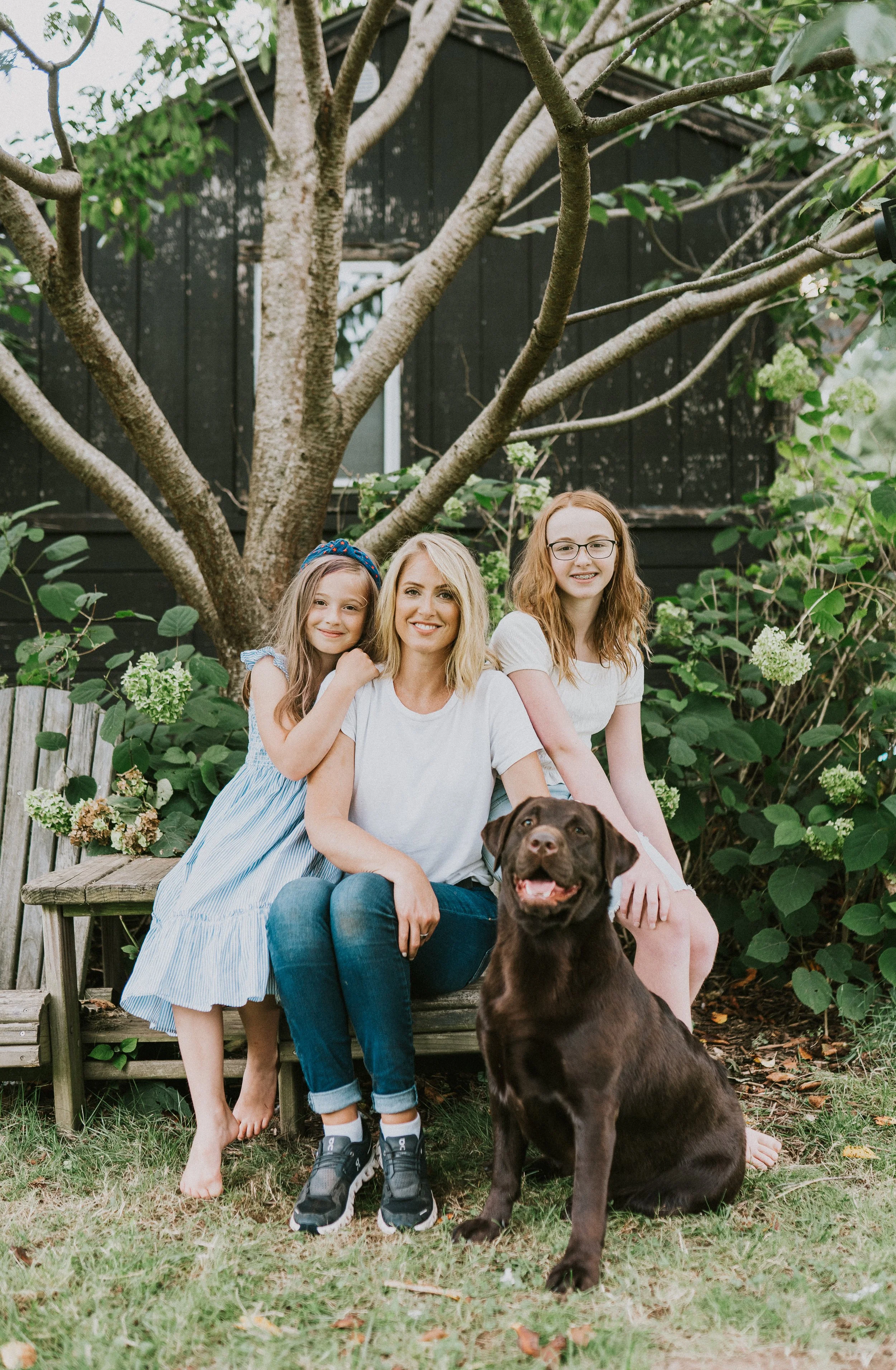 A woman with two young girls and a chocolate Labrador outside in a garden with a large tree and green plants in the background. The girl on the left is hugging the woman, and the girl on the right is sitting next to her. The woman and the girls are s