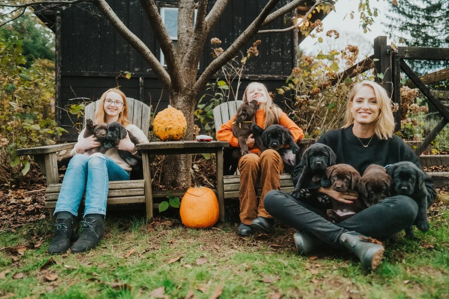 CopperRidge family sitting on wooden benches outdoors surrounded by trees and plants, holding multiple black and brown Labrador puppies