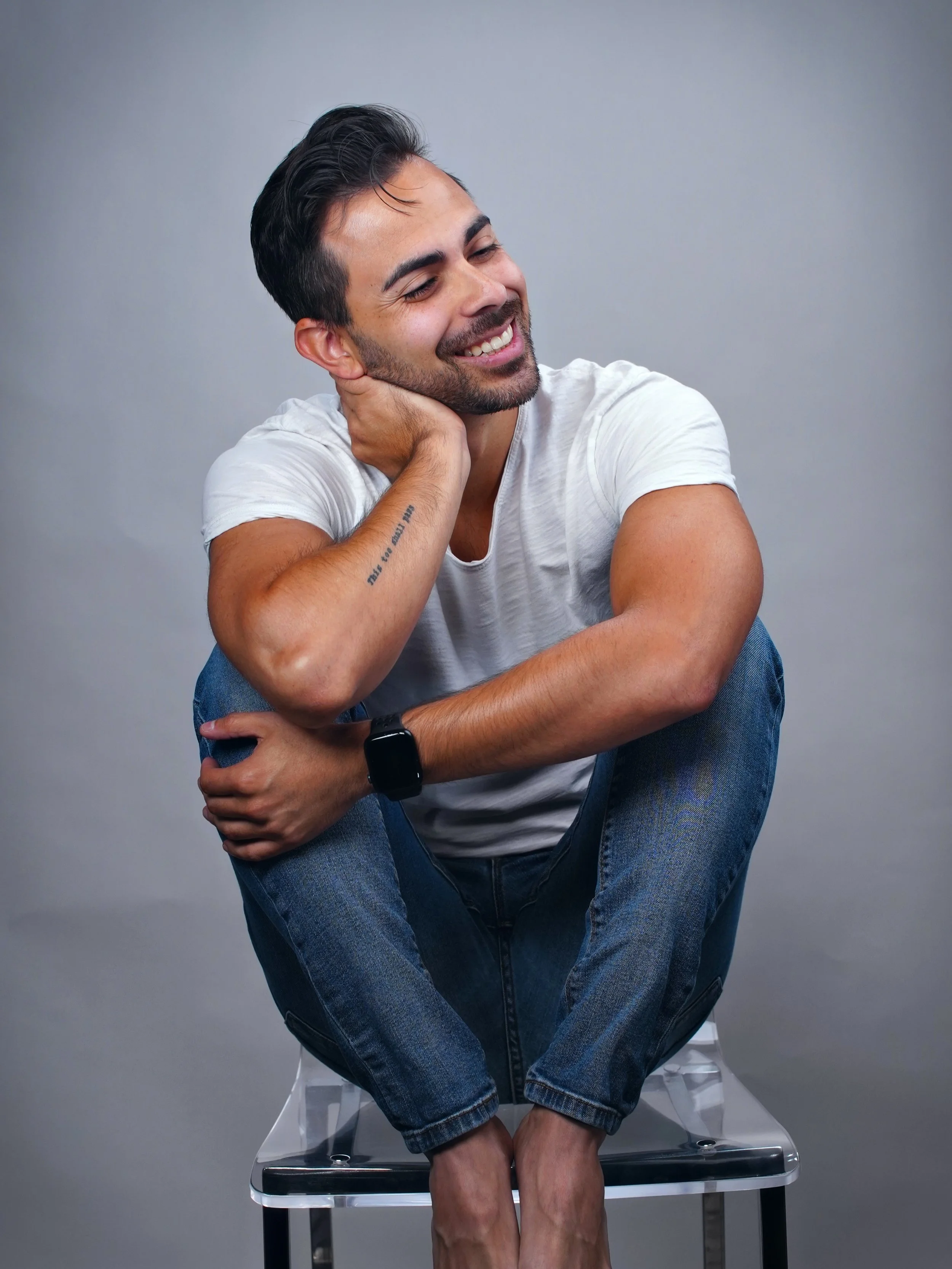 Man sitting on transparent chair in white t-shirt and jeans: Danny Morales — Mirror Method coach for Gay and Bisexual men