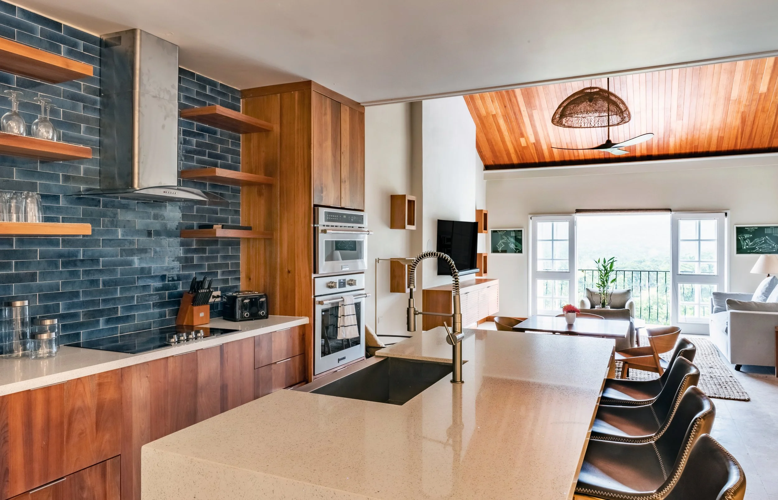 Modern open-concept kitchen and living room with wood accents, black subway tile backsplash, and large windows with a view of greenery.