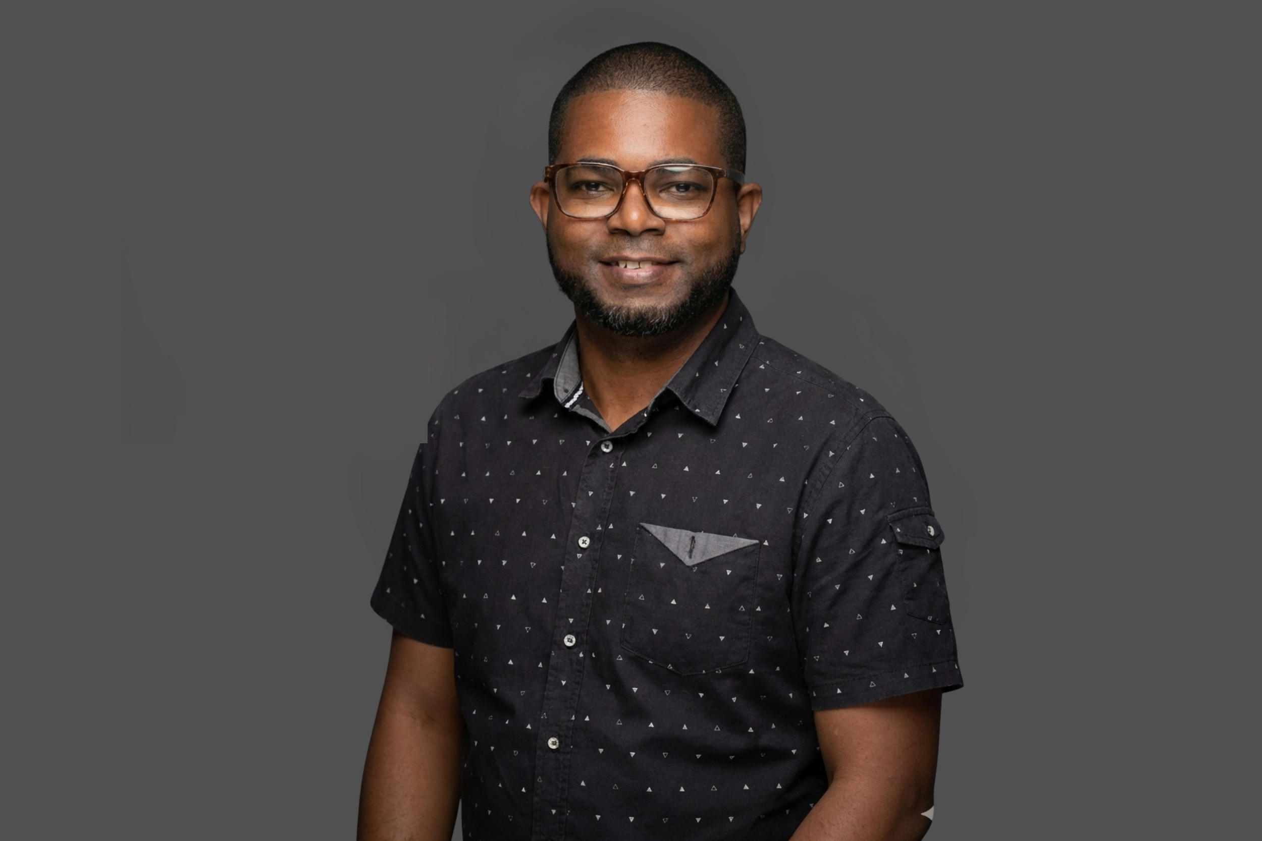 Portrait of a man with glasses, short beard, wearing a black patterned shirt, smiling, against a grey background.