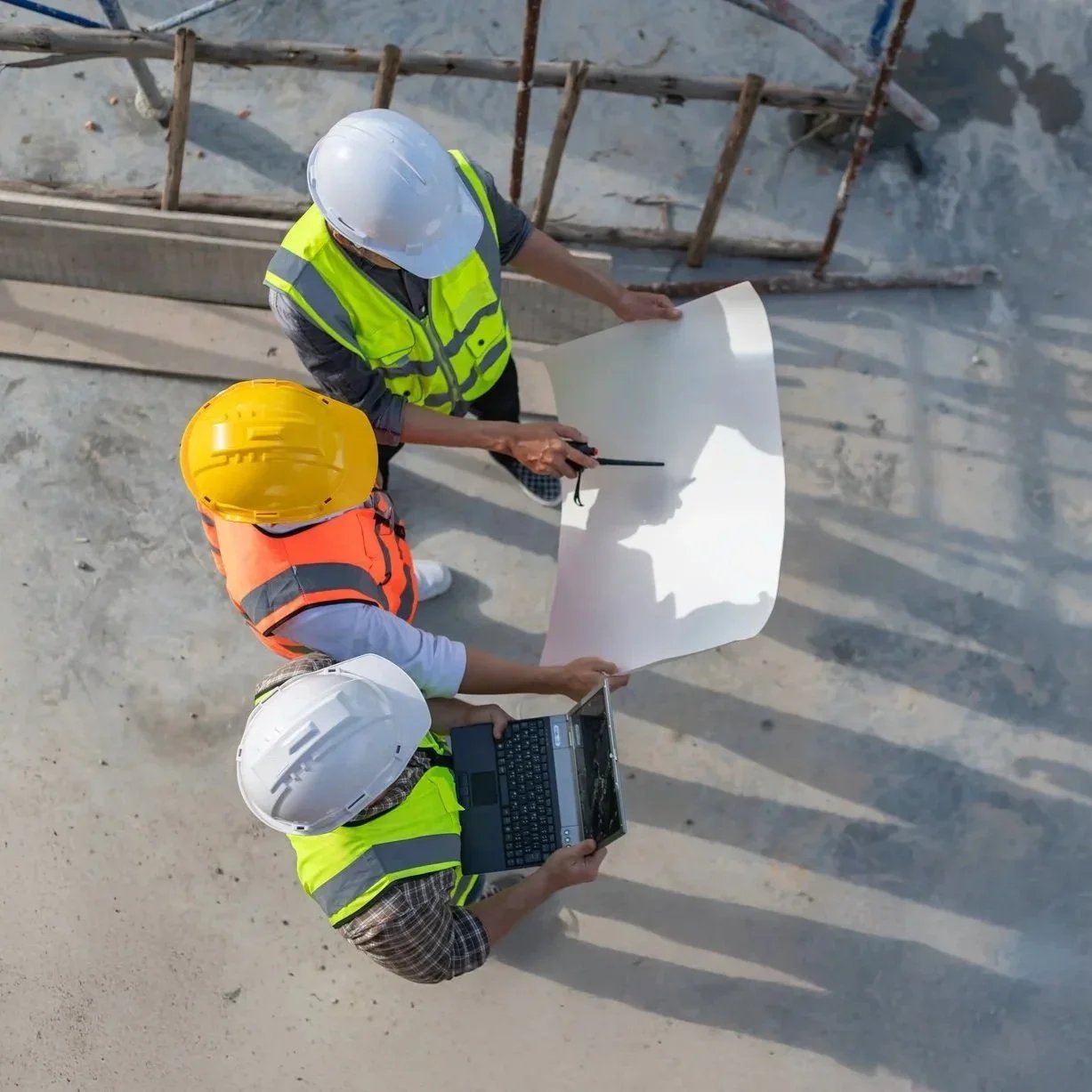 Three construction workers wearing safety helmets and reflective vests reviewing blueprints at a construction site.