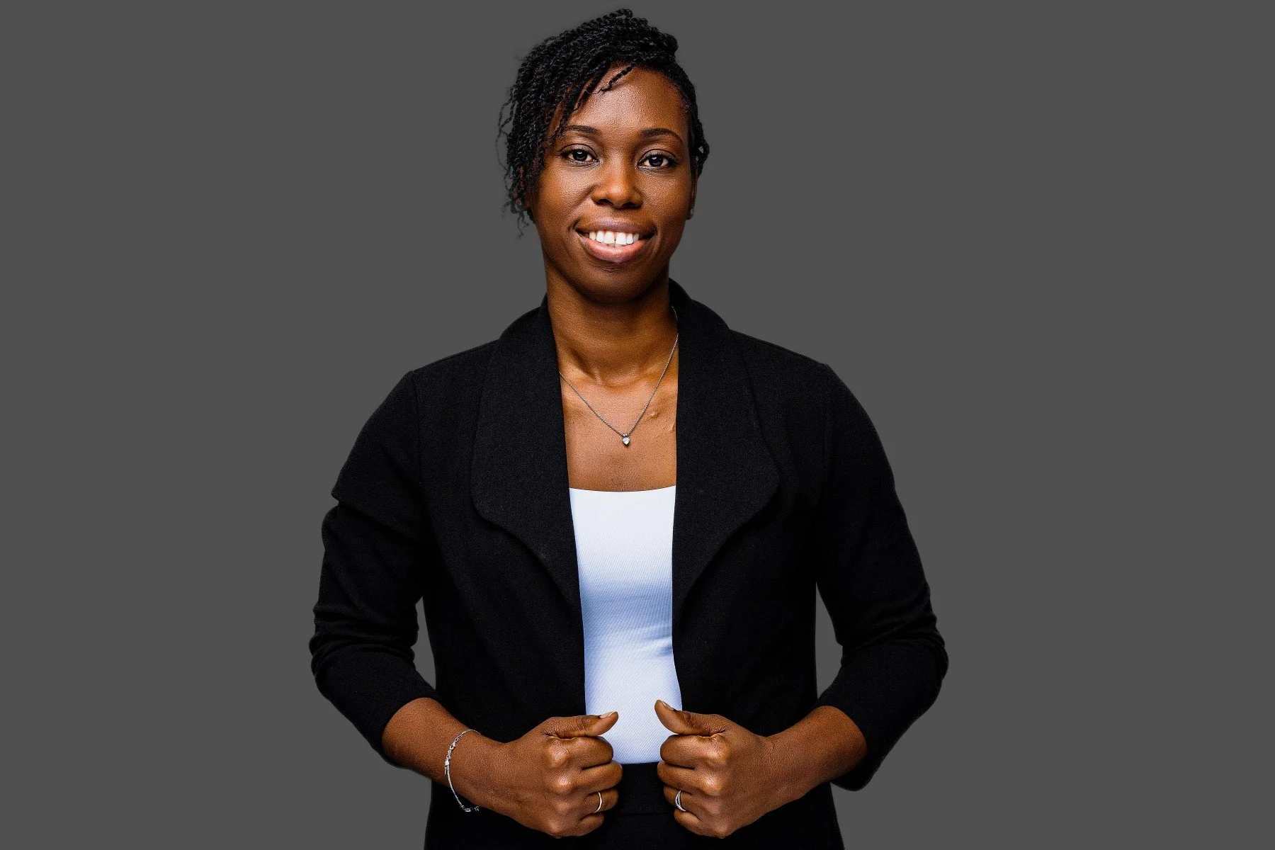 A professional woman with dark skin, wearing a black blazer over a white top, smiling, and posing against a gray background.
