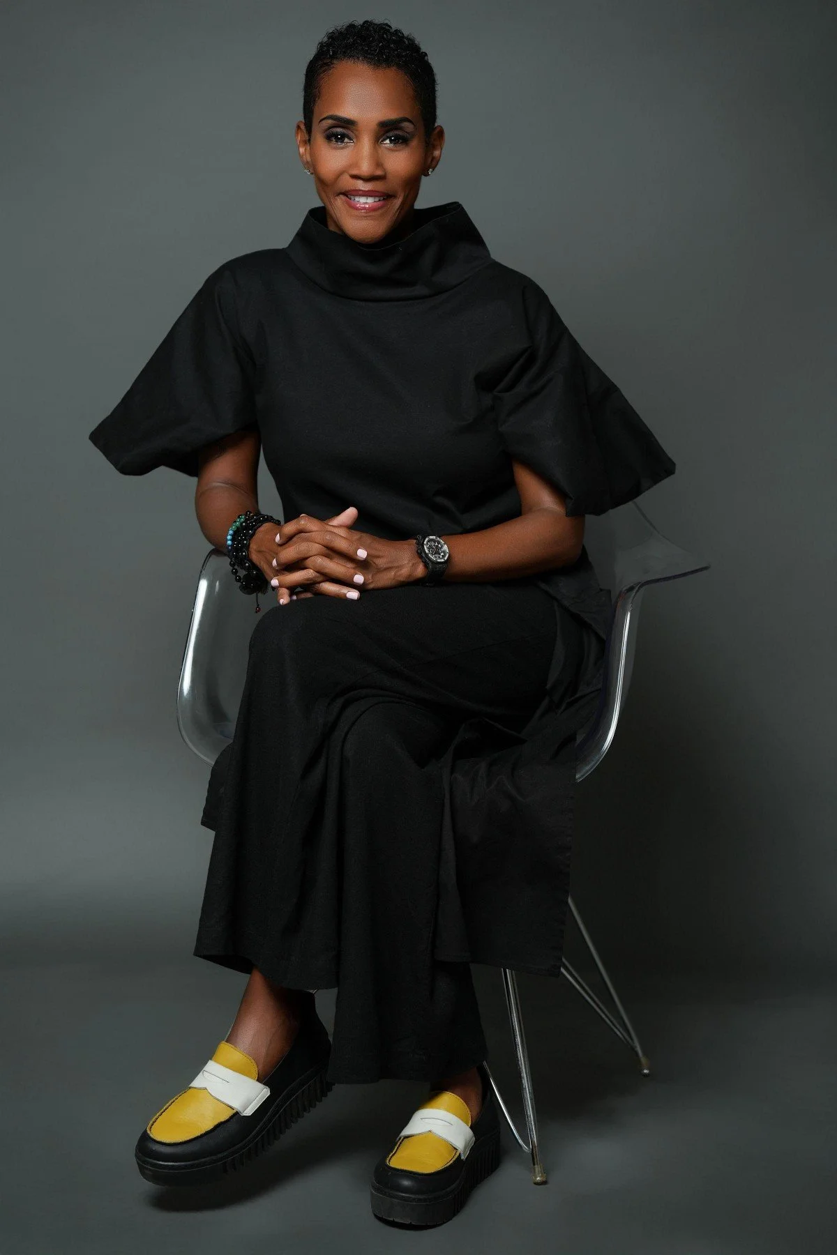 A woman with short black hair and dark skin, wearing a black high-collared top and black wide-leg pants, seated on a metallic chair against a gray background. She is smiling, with her hands clasped on her lap, wearing a watch and multiple bracelets, and sporting yellow and black chunky shoes with white straps.