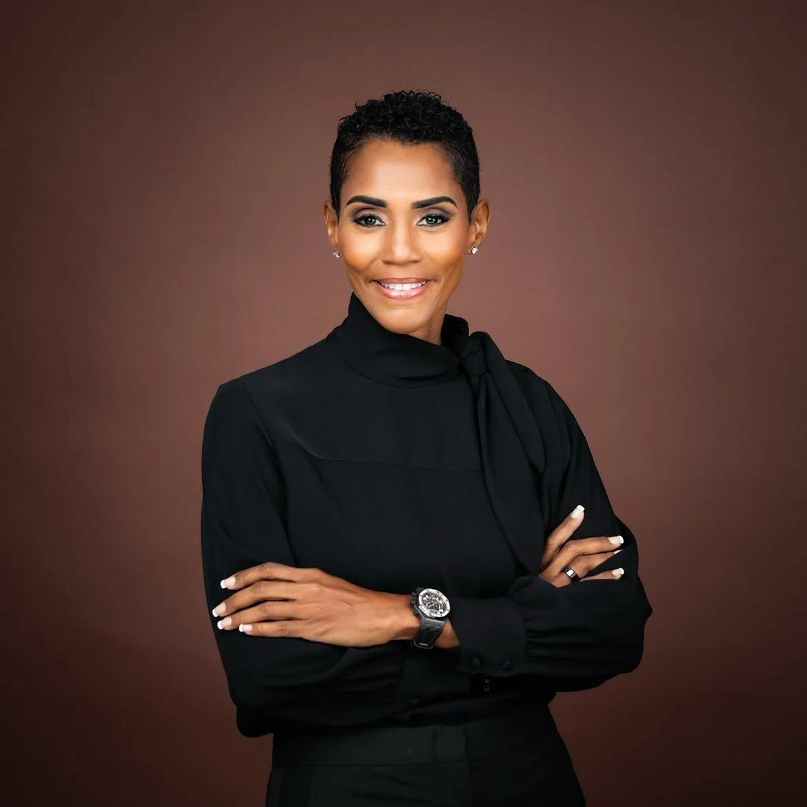 Professional woman (Isiaa Madden) with short curly hair wearing a black blouse with a bow at the neck, standing against a brown background, smiling with arms crossed.