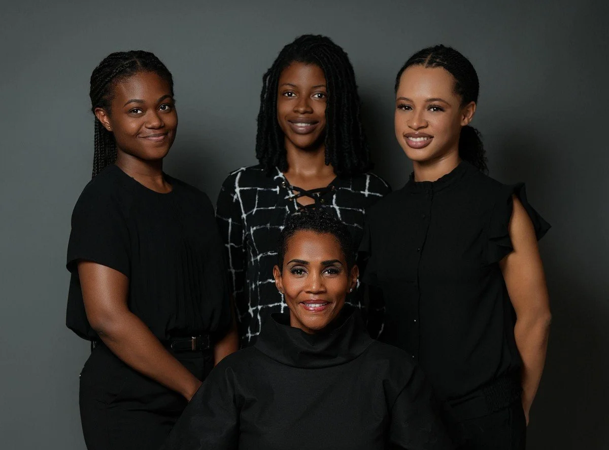 Four women on Design HQ's team standing in front of a gray background, smiling, dressed in dark-colored clothing.