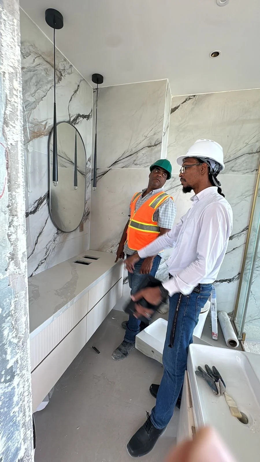 Two construction workers wearing hard hats and safety vests talking in a modern bathroom under construction, with marble walls, a mirror, and construction tools.