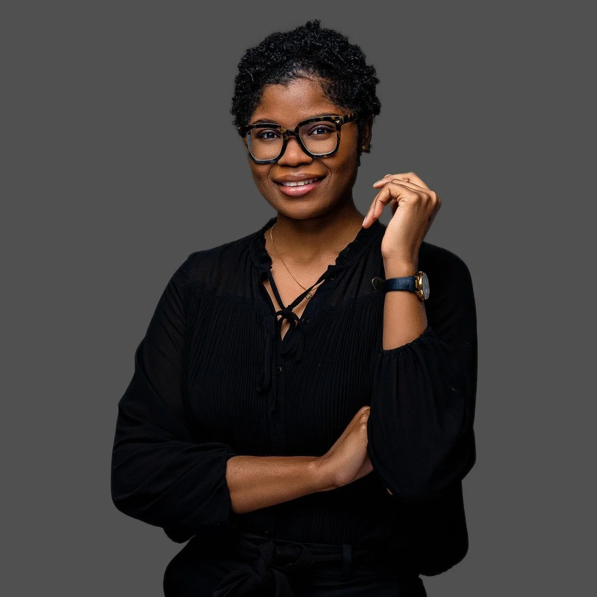 Portrait of a smiling Black woman with glasses, wearing a black blouse, a watch, and a necklace, posing with her arm crossed against a gray background.