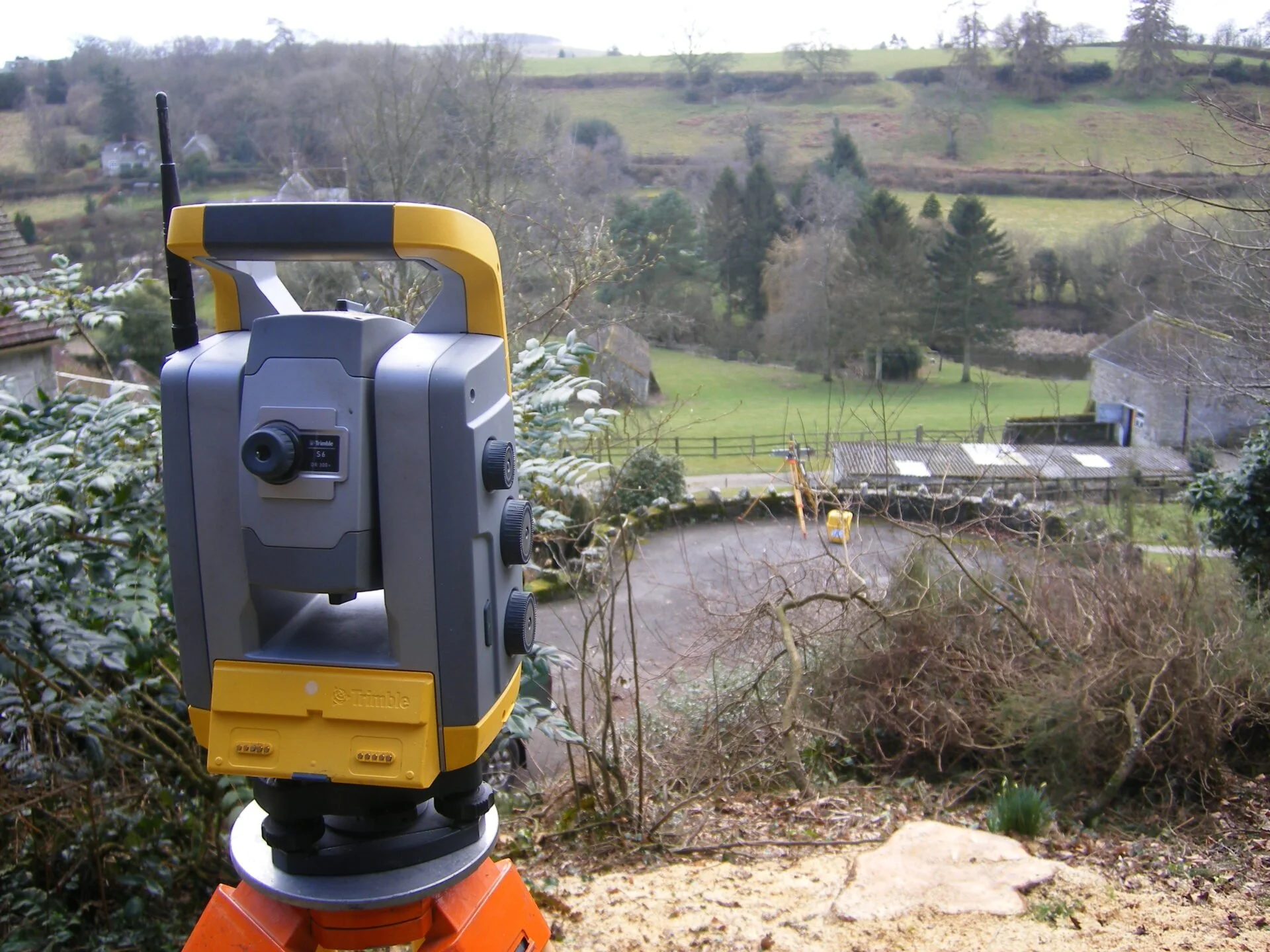 A yellow and gray laser level mounted on a tripod outdoors with a rural landscape in the background, including trees, houses, and rolling hills.