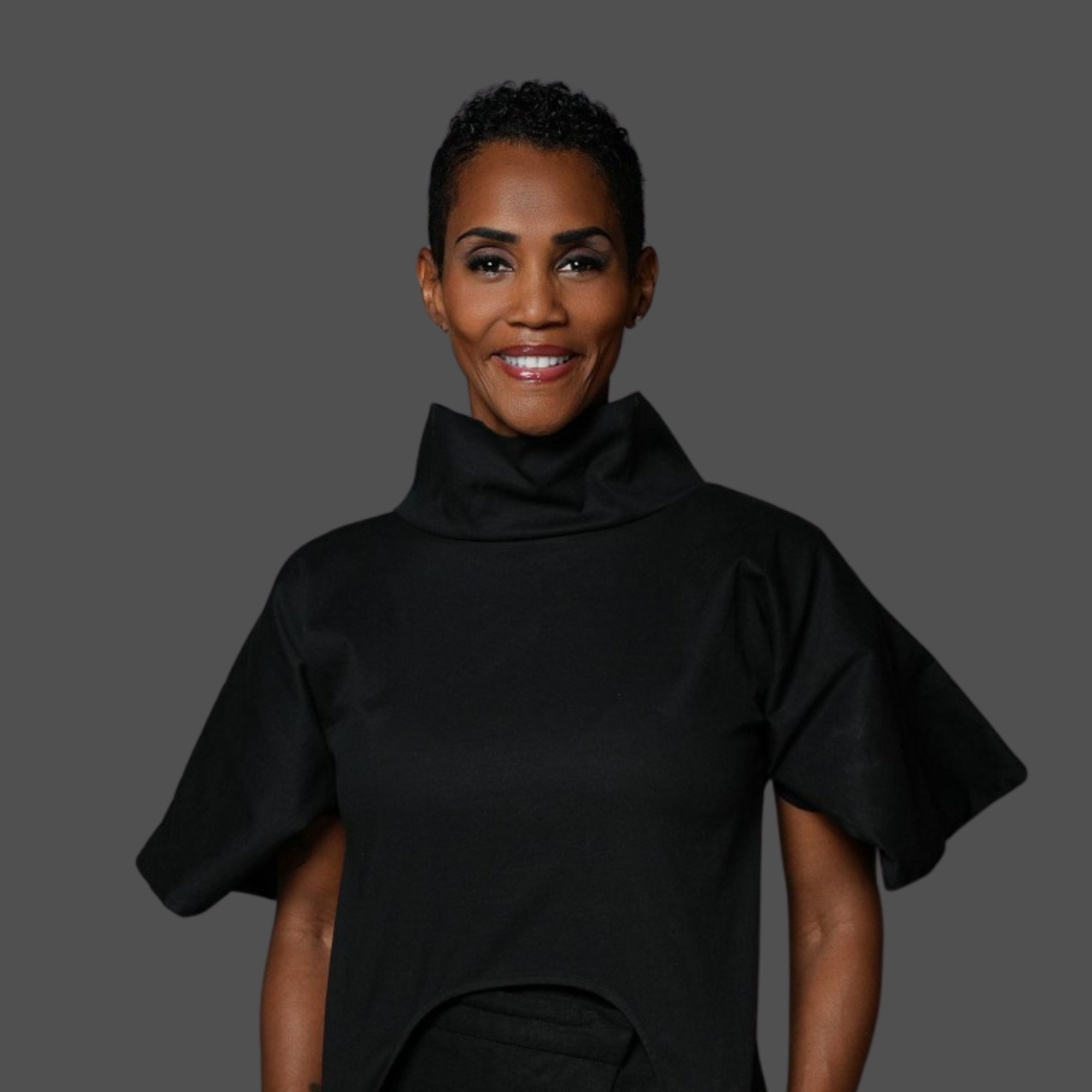 A woman (Isiaa Madden) with short curly hair, wearing a black turtleneck, is smiling at the camera. The background is gray.