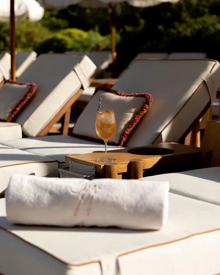 Poolside lounge chairs with cushions, a rolled towel on a table, and a cocktail glass with an orange drink garnished with an orange slice and a cherry, in an outdoor setting.