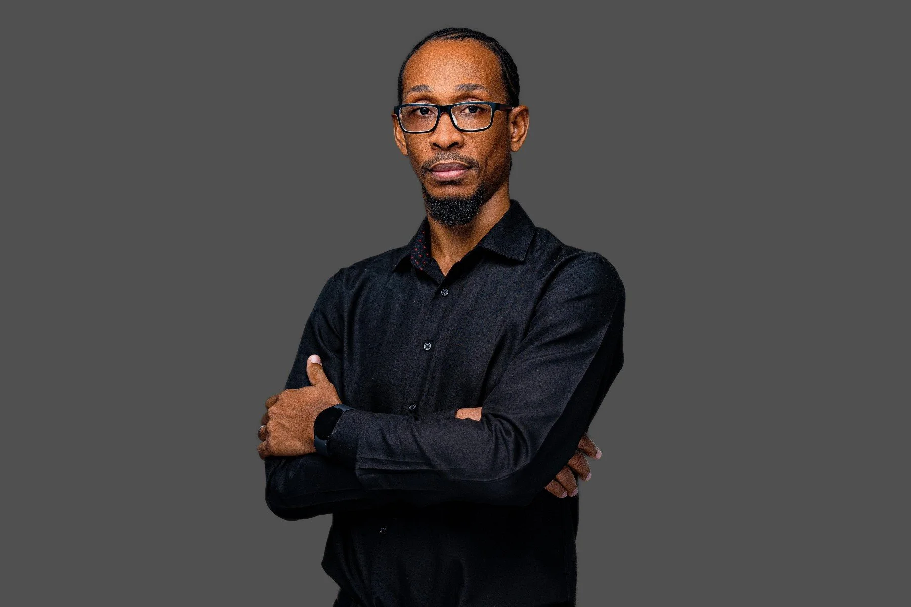 A man with glasses, a goatee, wearing a black button-up shirt, standing with arms crossed against a dark gray background.