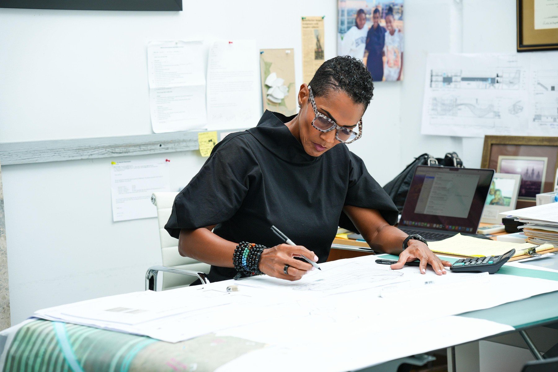 A woman (Isiaa Madden) with short curly hair and glasses sits at a cluttered desk in an office, drawing on large sheets of paper with a pen, surrounded by office supplies, electronic devices, and photos on the wall.