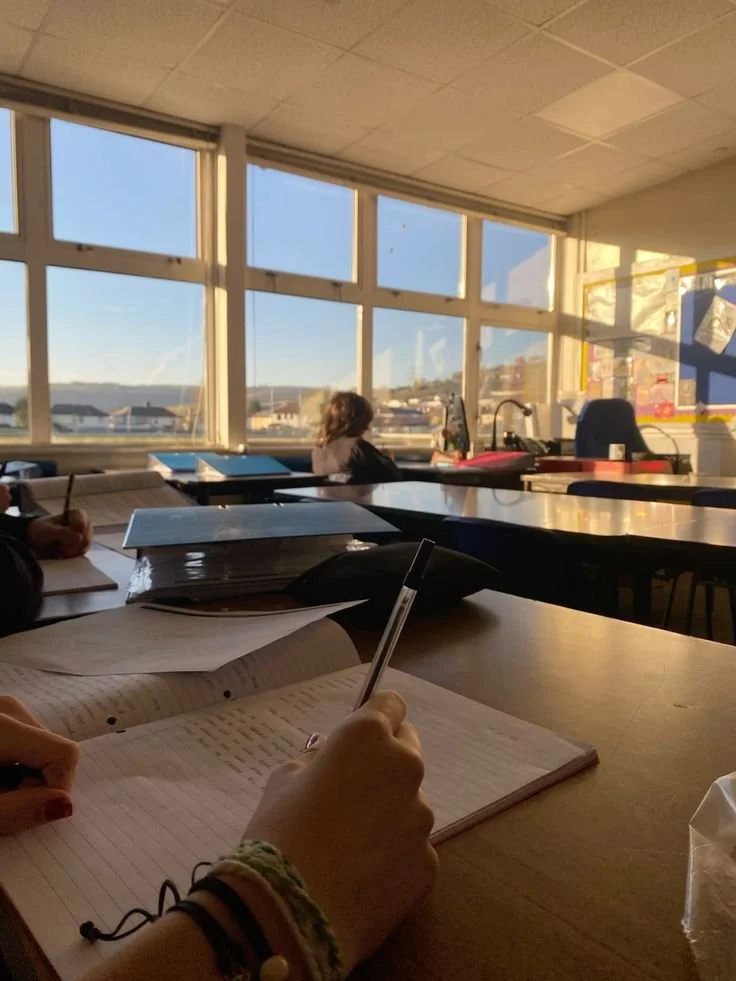 A classroom with sunlight coming through large windows, students working at their desks, one holding a pen and writing in a notebook.