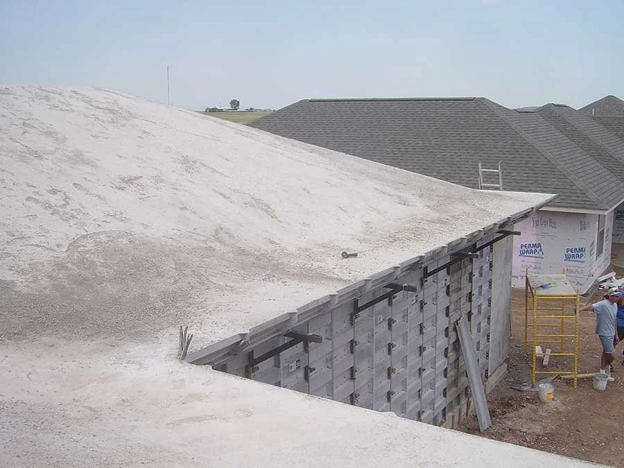 Construction site of a house with a sloped roof, workers, and building materials visible, with neighboring houses in the background.