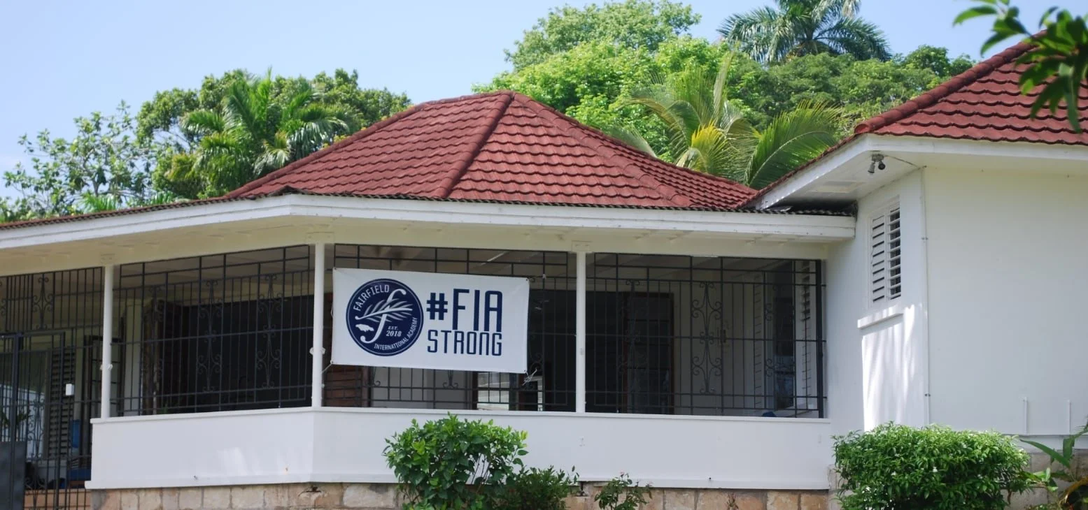 A school with a red tiled roof, white walls, and a front porch with a sign that reads '#FIA STRONG' and features a logo with a wave and the words 'Fairfield International'. There is lush green foliage and trees in the background.