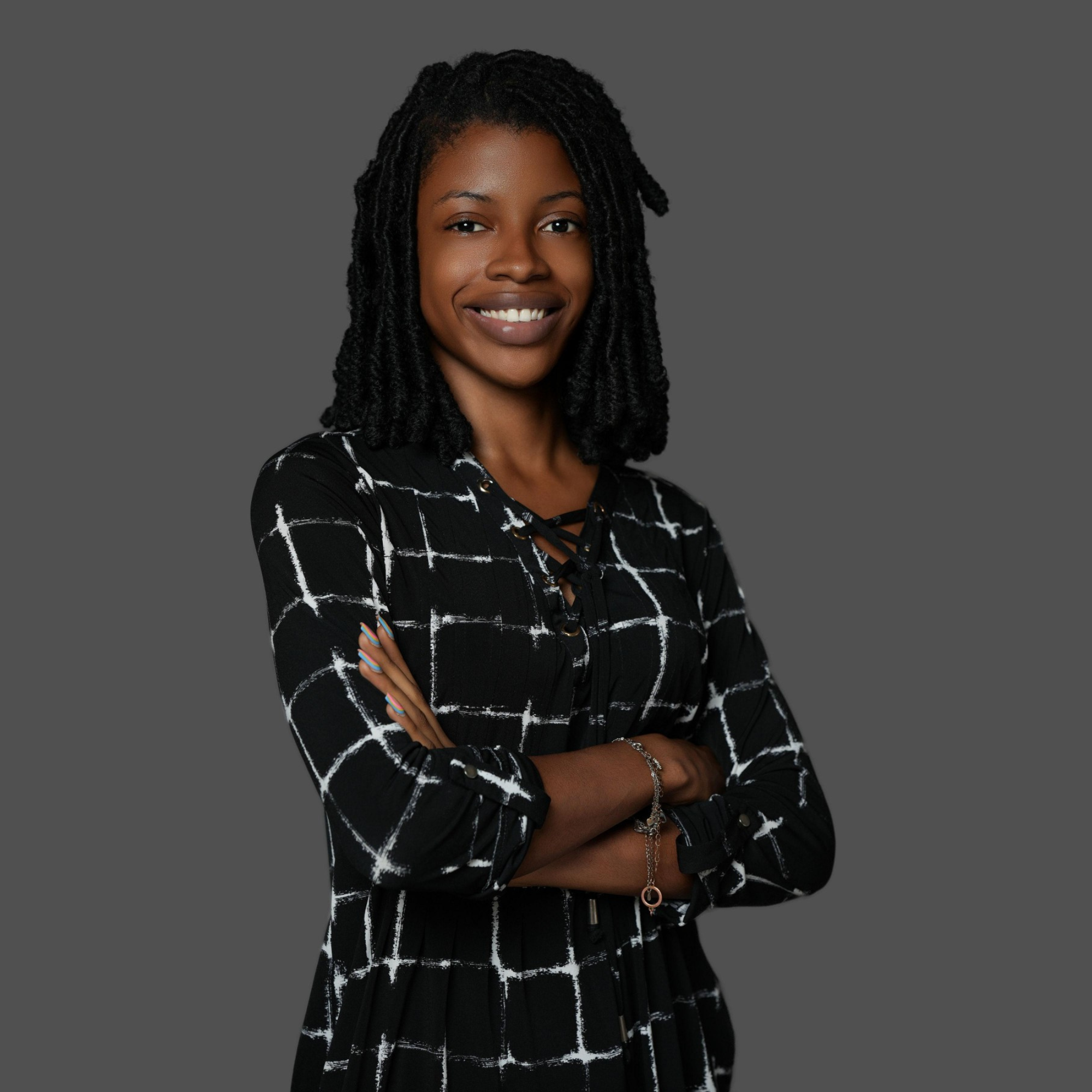 Portrait of a smiling woman with locs, wearing a black and white patterned top. She has her arms crossed and is standing against a gray background.