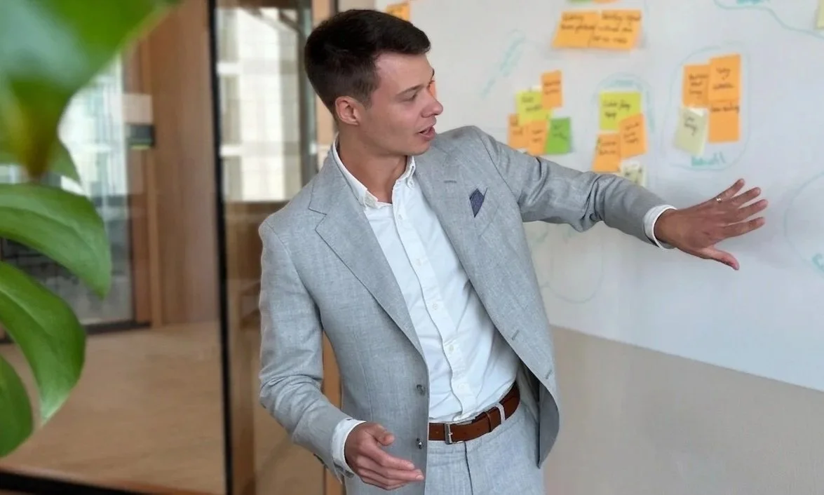 Ein Mann in einem grauen Anzug erklärt auf einer Whiteboard-Tafel mit bunten Haftnotizen.
