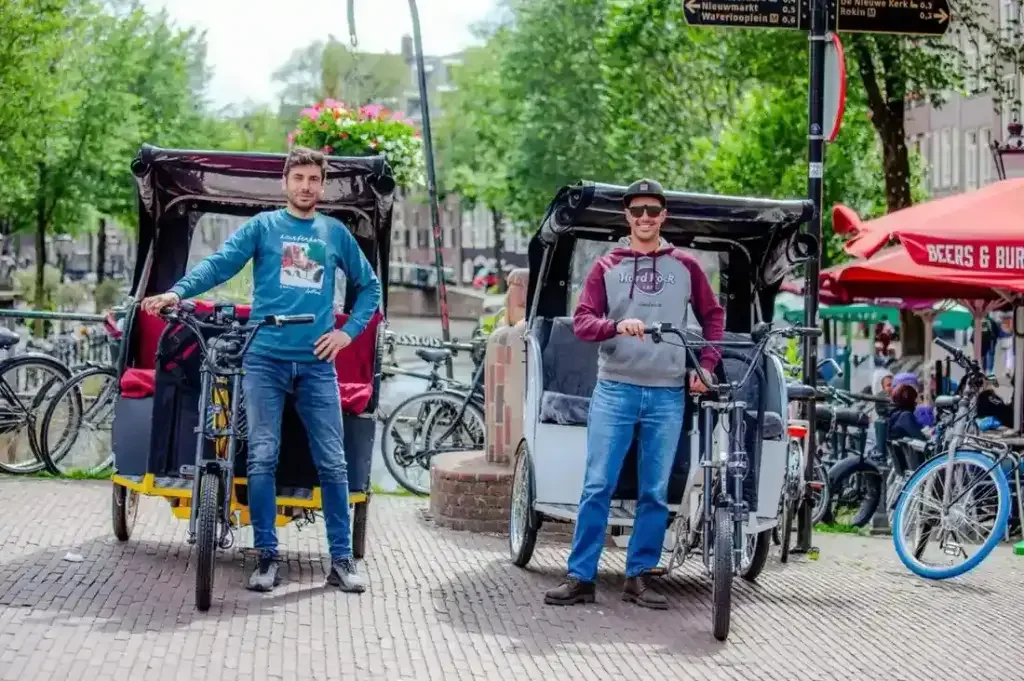 Two men standing with their pedicabs in an urban setting, surrounded by bicycles and outdoor seating, with trees and buildings in the background.