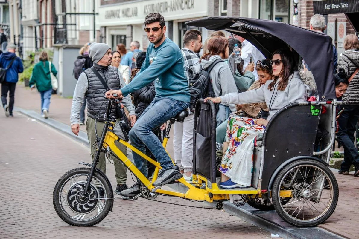 A rickshaw with a man riding a bicycle with a passenger seat in the back, carrying multiple women. The park is crowded with pedestrians.
