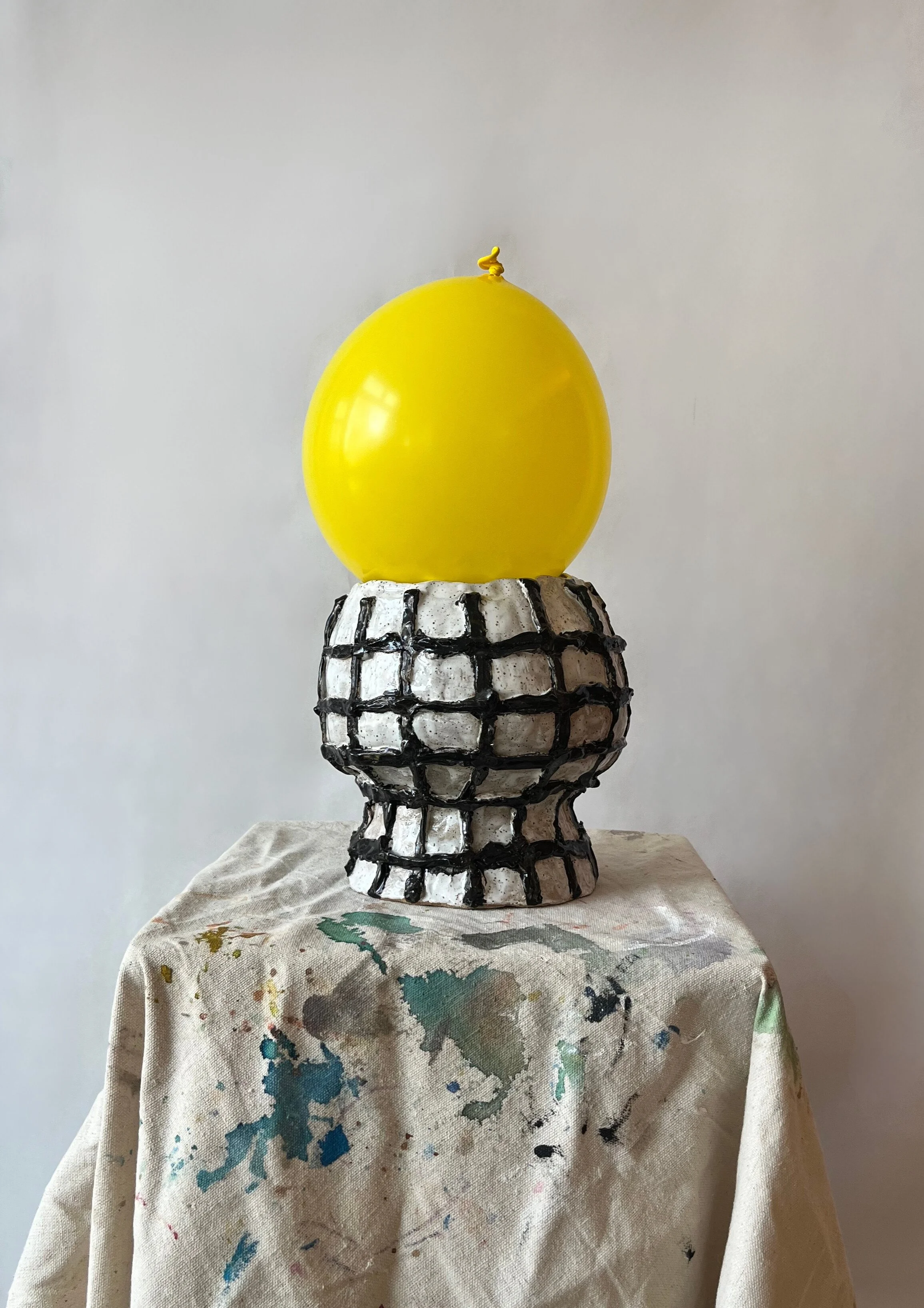 A yellow balloon on top of a black and white ceramic sculpture resembling a pineapple, placed on a cloth-covered surface.