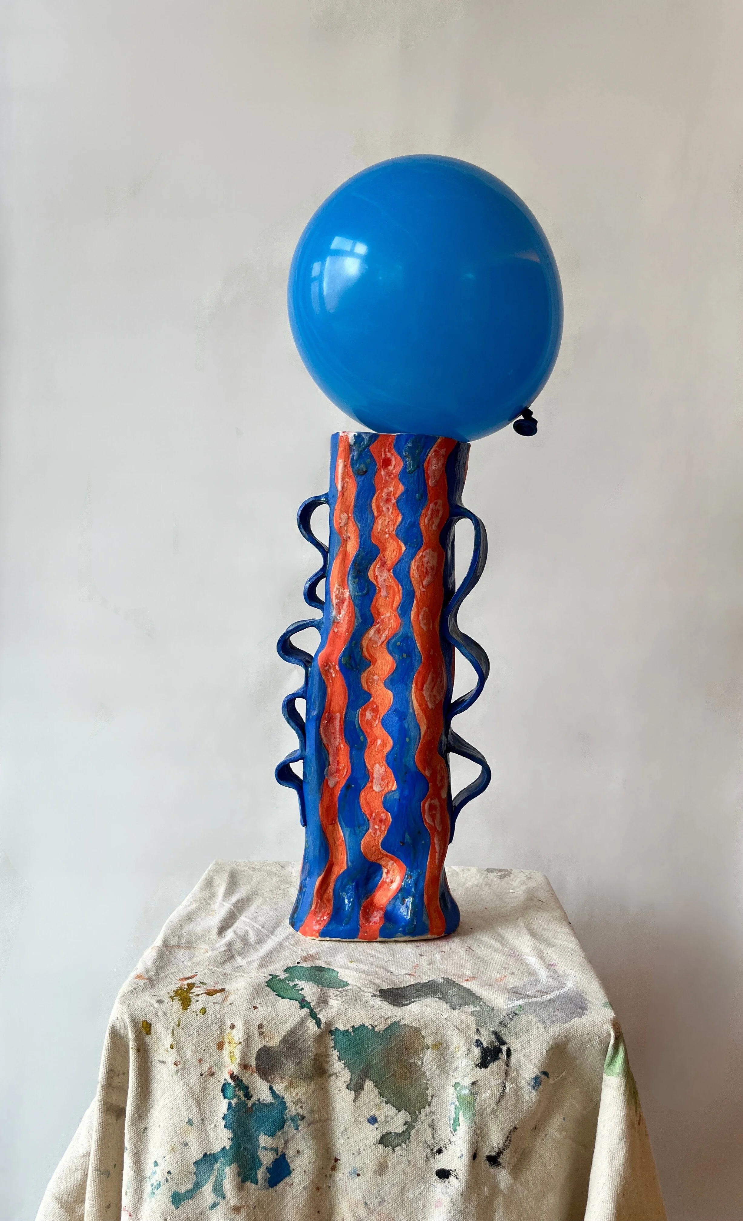 A colorful ceramic sculpture resembling a jellyfish, with a blue balloon-like top, a tall wavy neck painted with wavy orange, blue, and green lines, and long dangling tentacles, placed on a paint-stained cloth.