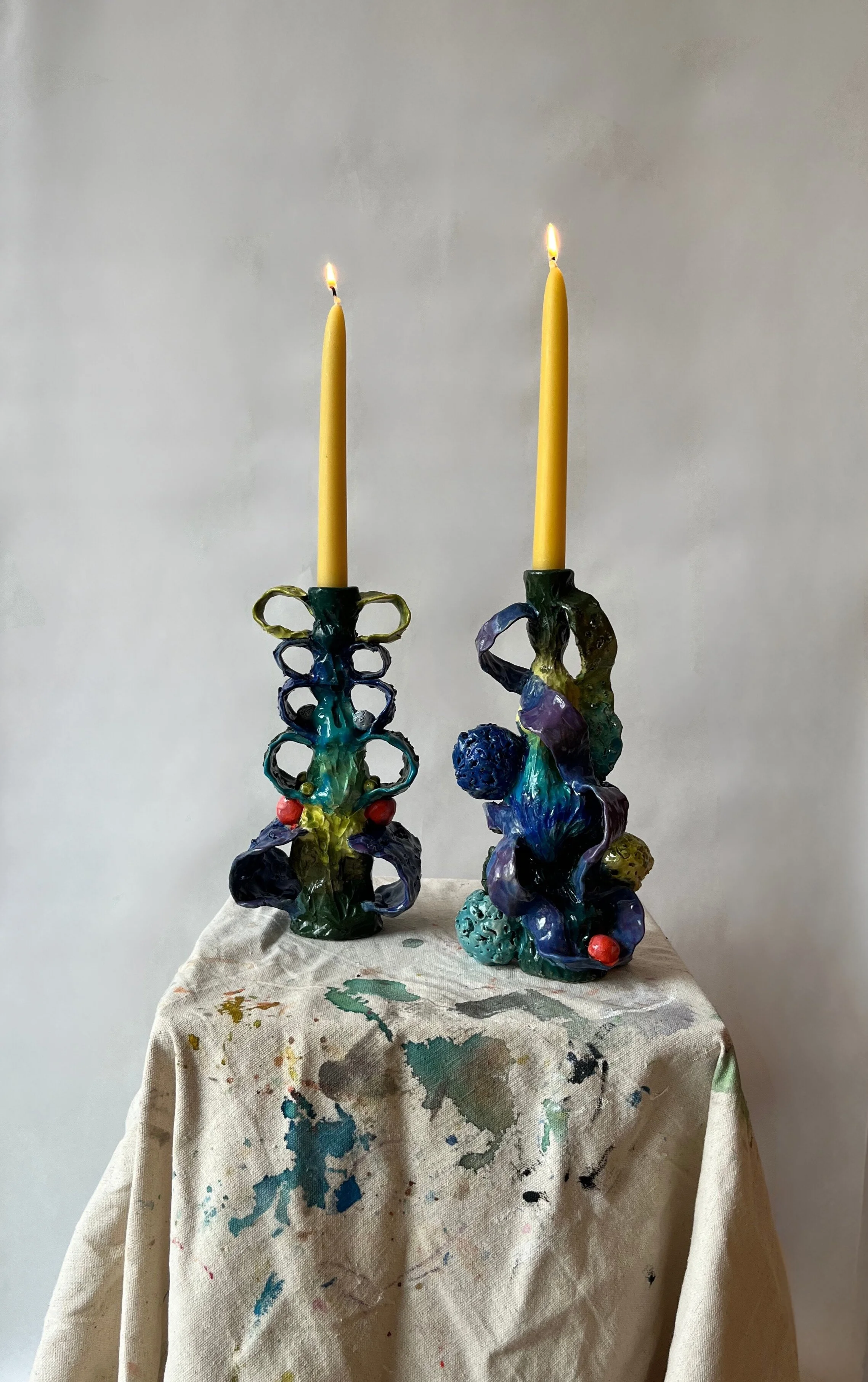 Two colorful ceramic candlestick holders with yellow taper candles, standing on a paint-stained fabric-covered surface against a plain background.