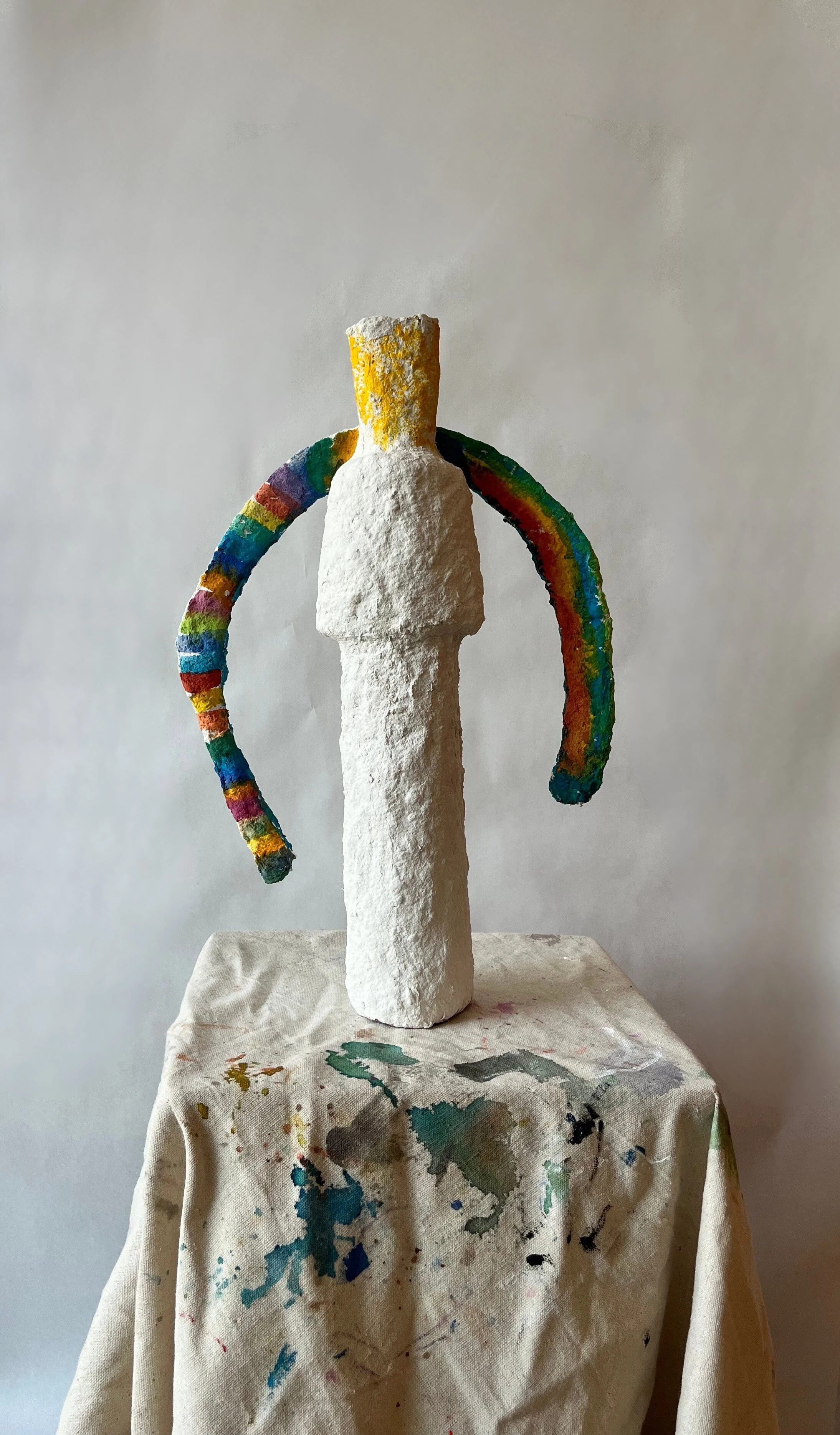 An abstract sculpture resembling a stylized human figure with a tall, white body and rainbow-colored, curved arms extending outward. The sculpture is on a painter's palette covered table with paint stains, against a plain, light background.