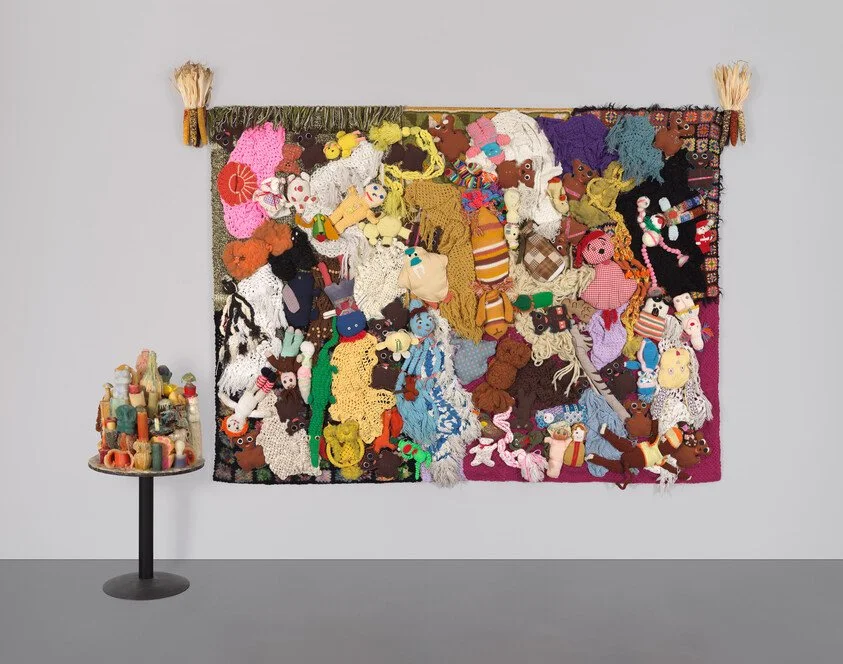 Colorful textile art with various stuffed animal figures, hanging on a wall. Small table with spools of thread beside it.