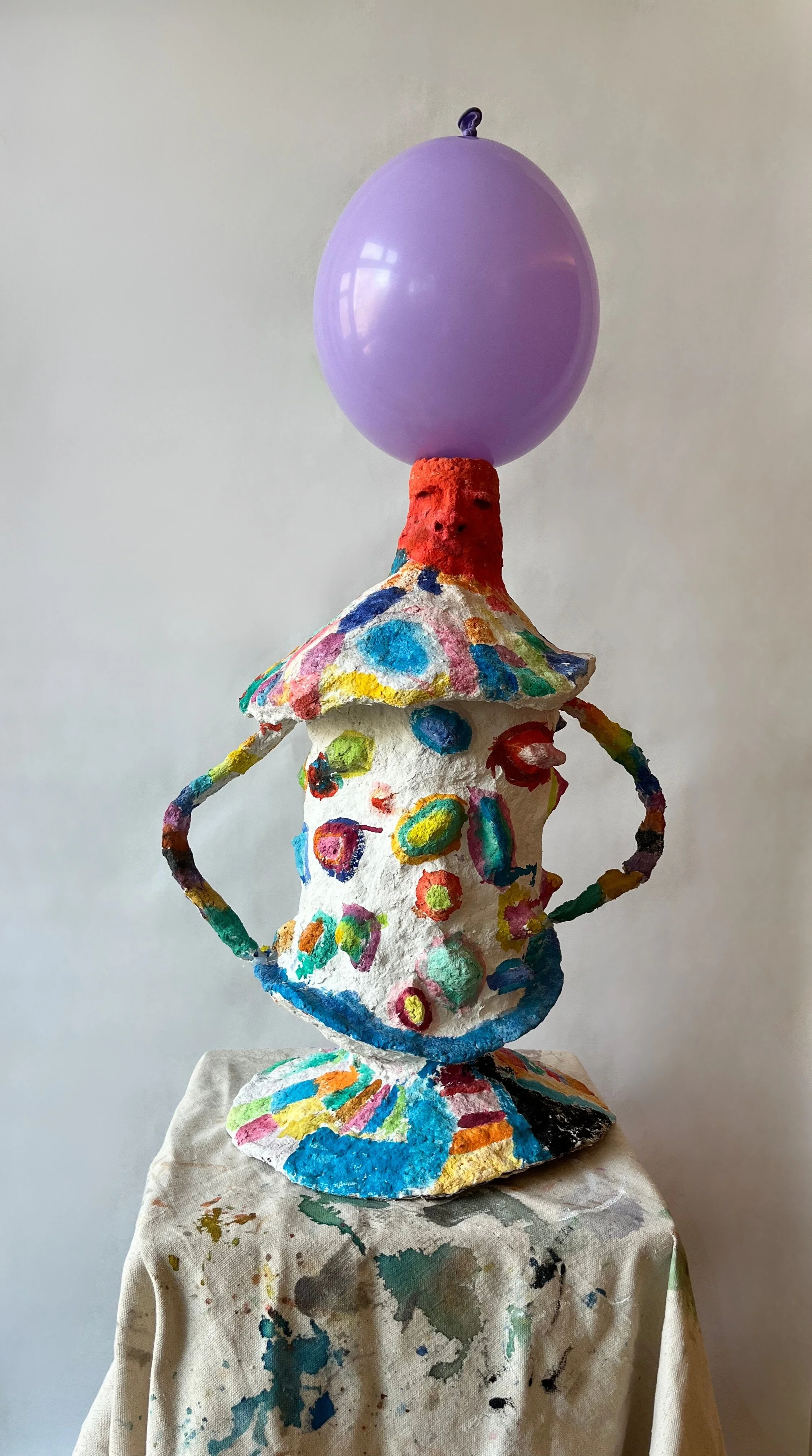 Colorful, abstract sculpture of a figure with a purple balloon head, painted with multicolored dots and shapes, standing on a decorated base.