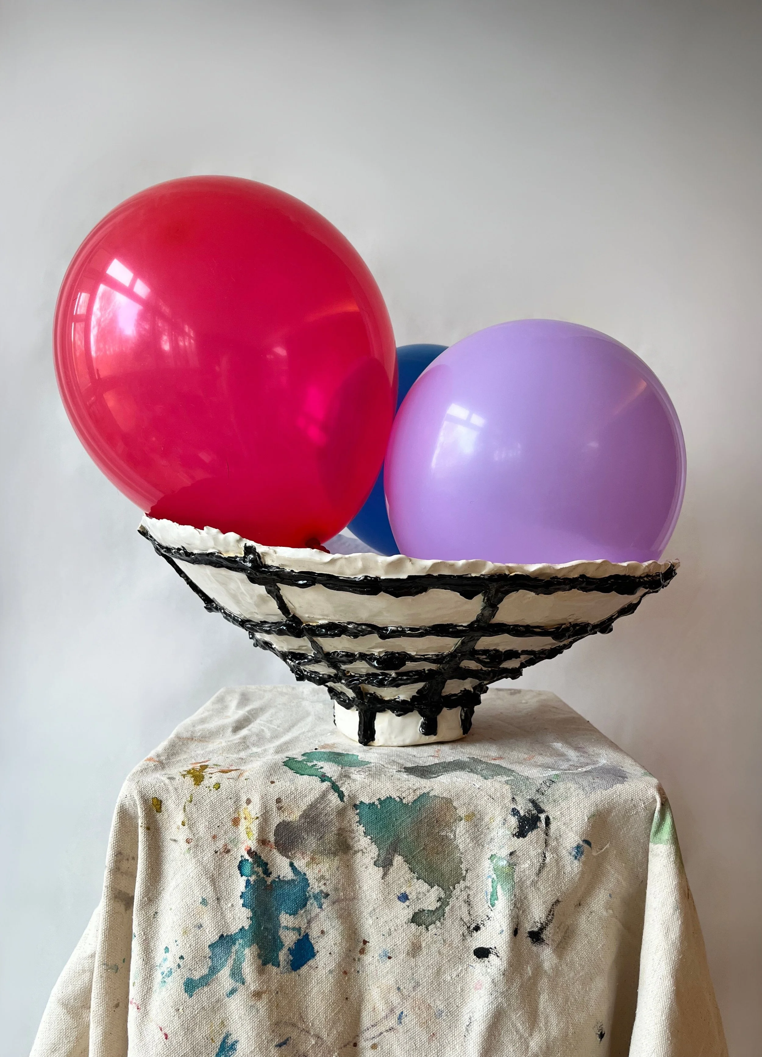 A ceramic bowl with a black frame holding three balloons, one red, one blue, and one purple, against a plain wall background, placed on a table covered with a paint-splattered cloth.