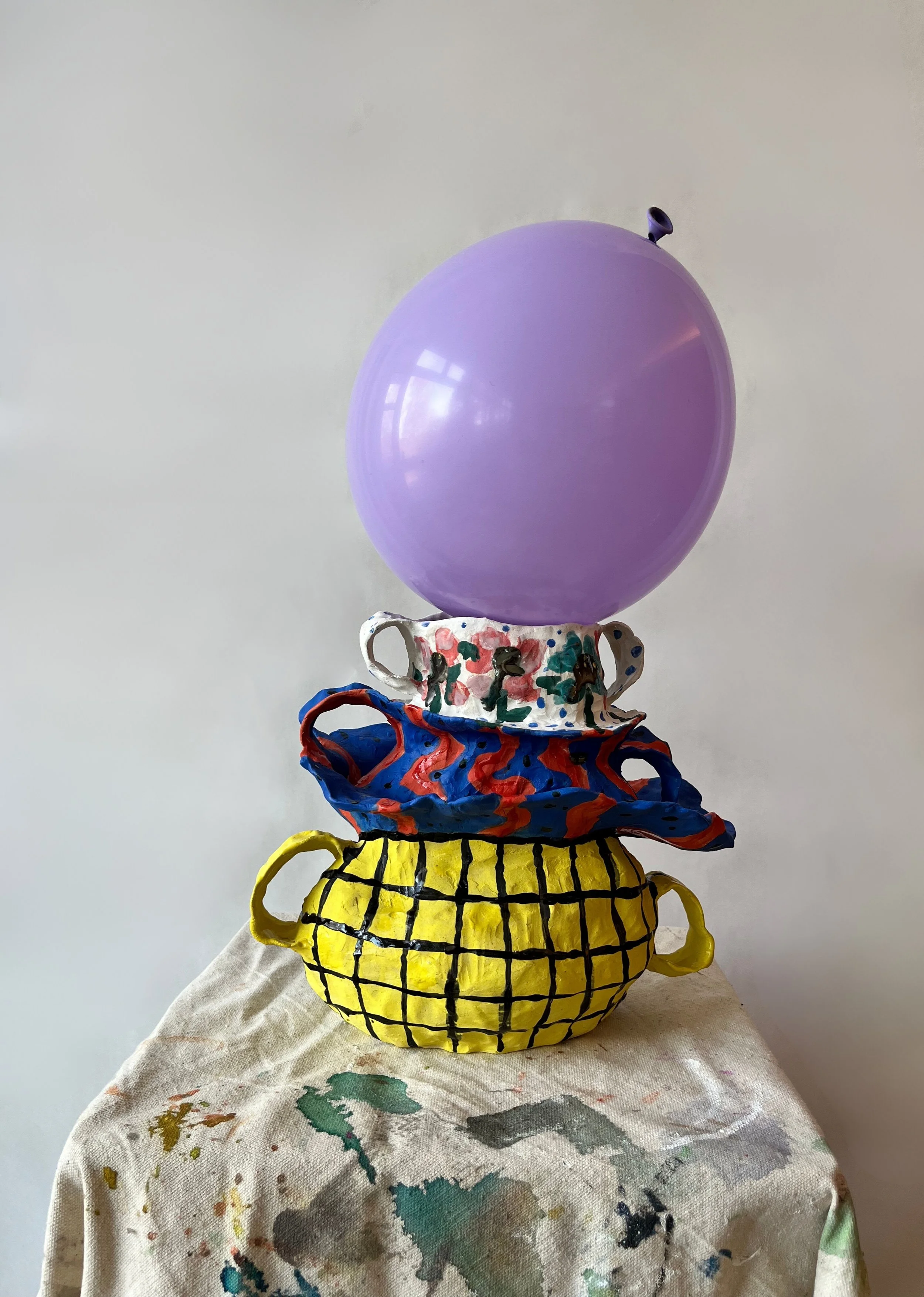 Colorful ceramic teapots stacked on top of each other with painted designs, placed on a fabric with paint splatters against a plain wall