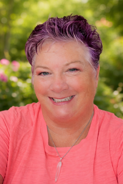 A smiling woman with short purple hair, wearing a pink top and a silver necklace, outdoors with green foliage in the background.