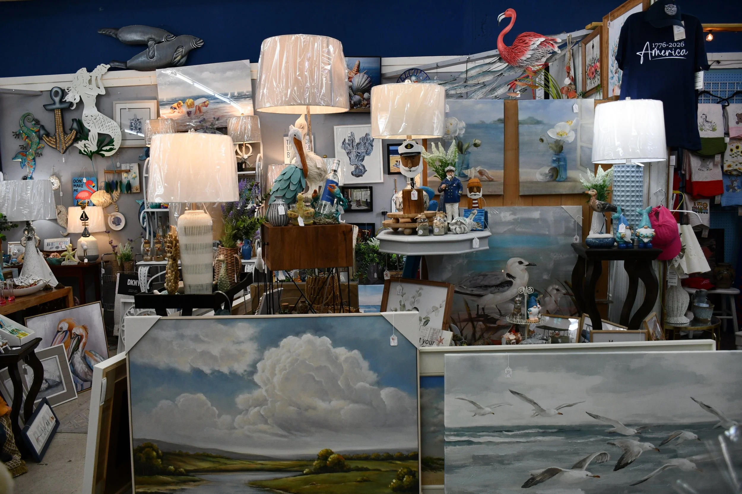 Interior of an art and home decor store with paintings of clouds, seagulls, and beaches, lamps, nautical themed decorations, and various decorative items.