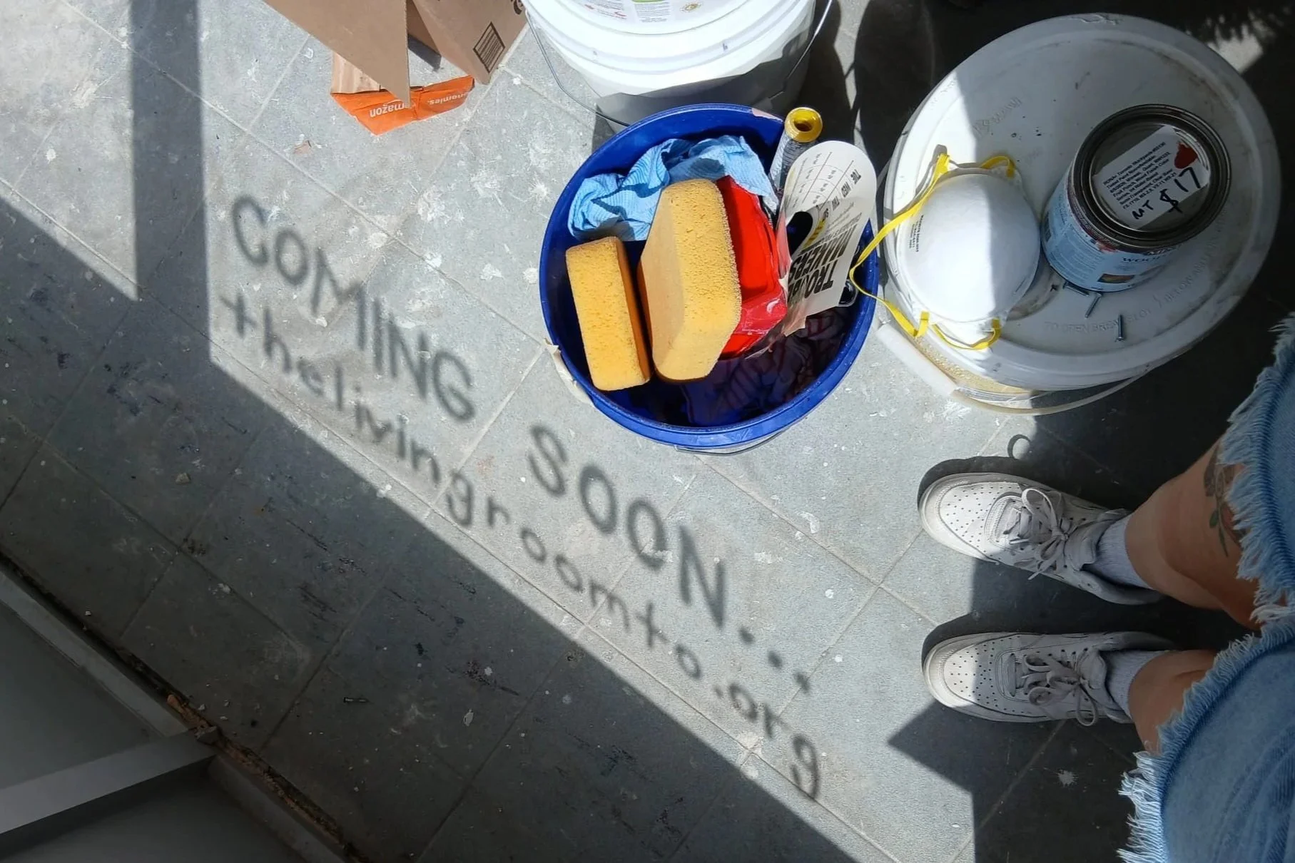 An outdoor scene showing a tiled patio with various painted shadows, including a faded shadow of a sign or text. There are two buckets, one containing cleaning sponges and rags, and the other has a white paint can with a yellow ribbon and a partially visible label. The image also shows a person's legs wearing white sneakers and denim shorts, standing on the patio.