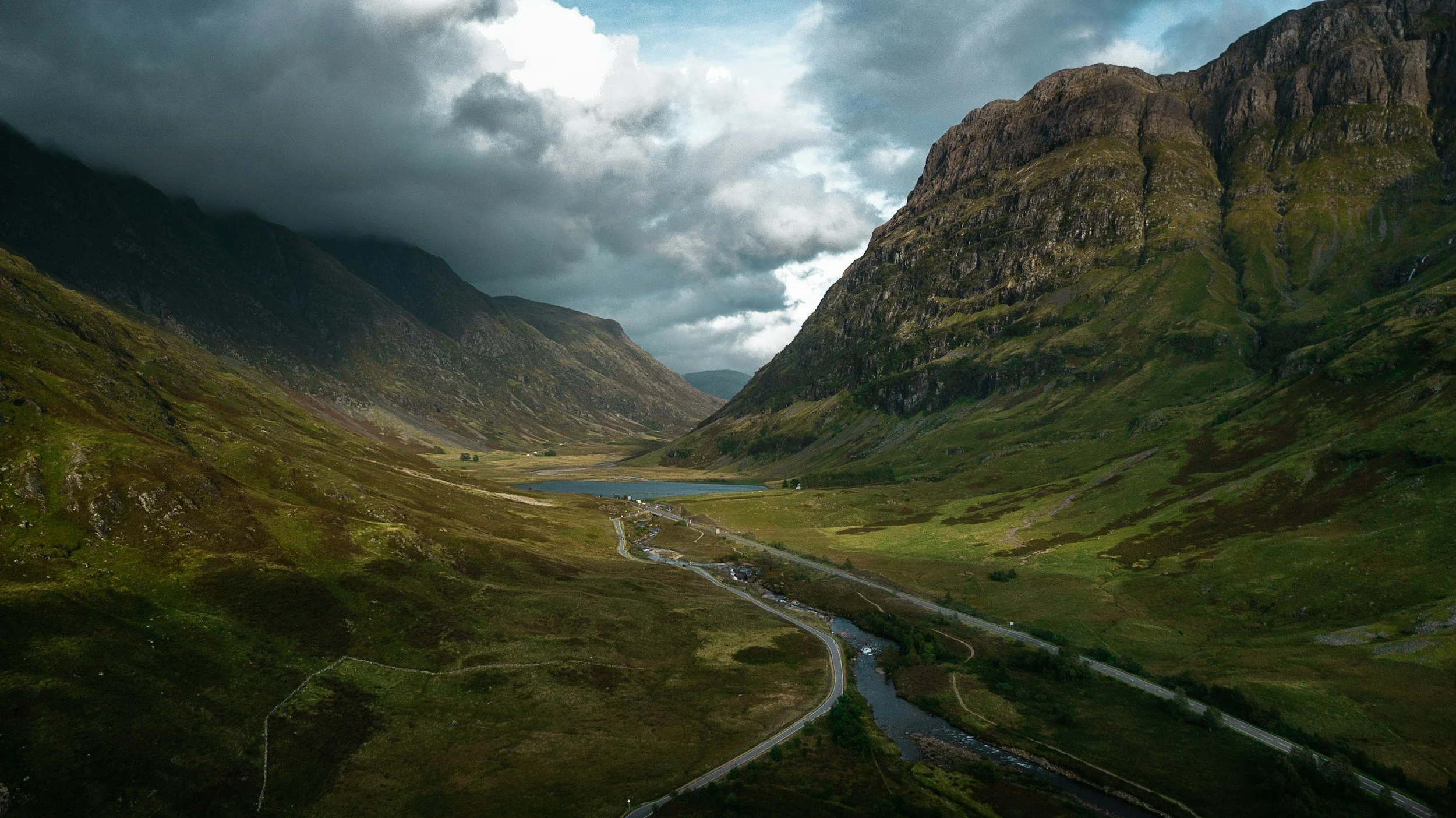 A scenic Scottish landscape of green mountains with a cloudy sky, a river flowing through the valley, and a small lake in the distance.