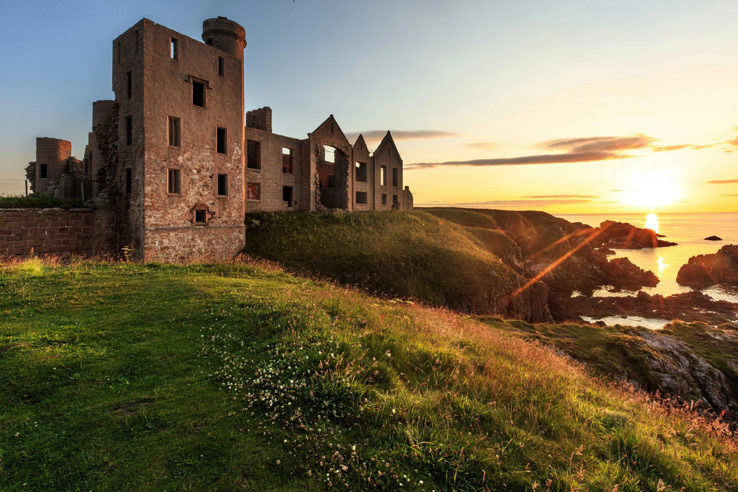 Ruined stone castle on a grassy hill overlooking the ocean at sunset, with clouds and water in the background.