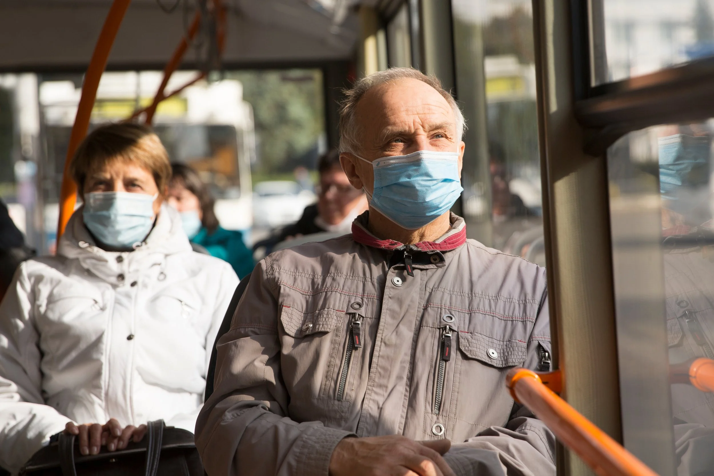 Two seniors wearing face masks riding on a bus, with sunlight coming through the windows.