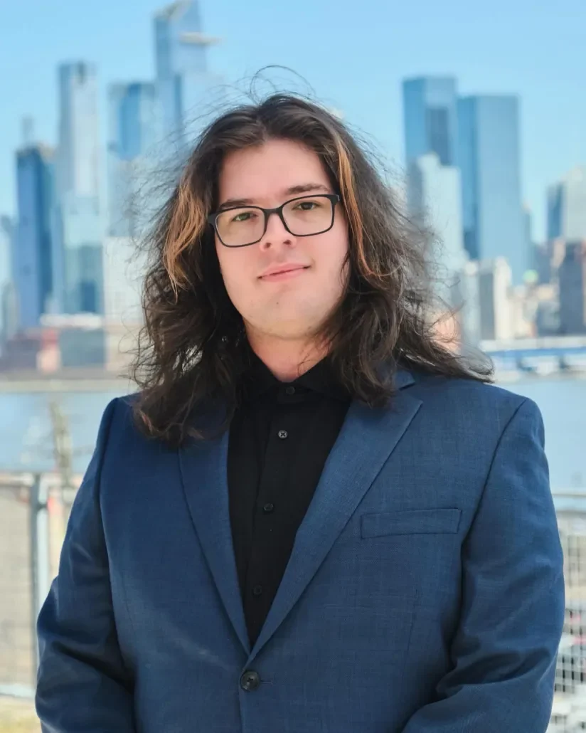 A young man with long, wavy brown hair, wearing glasses and a dark blue suit jacket, standing outdoors with a city skyline in the background.