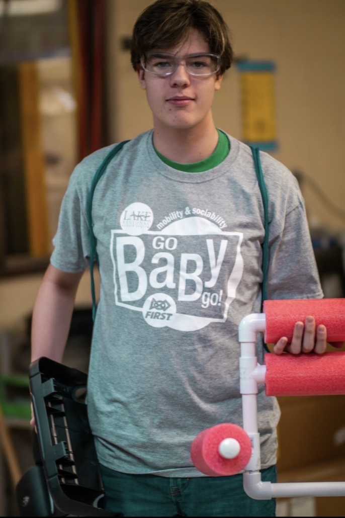 A young person with glasses, wearing a gray t-shirt that says "Go Baby Go!" and a green collar, stands indoors holding a pink foam roller attached to a white pipe structure. The background features shelves and a yellow board.