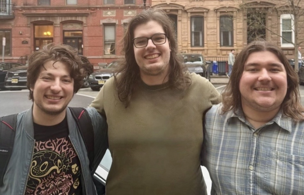 Three young men with long hair smiling and standing close together outdoors on a city street with brownstone buildings and parked cars in the background.