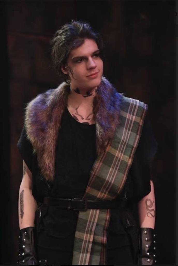 A young person with dark hair styled in a messy fashion, wearing a black outfit with a plaid sash. They have a faux fur collar, tattoos on their arms, and are standing in a dimly lit environment, possibly on stage.