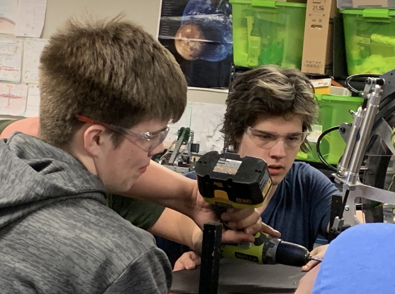 Two young men wearing safety glasses working together on a robotics project, with green storage bins and galaxy poster in the background.
