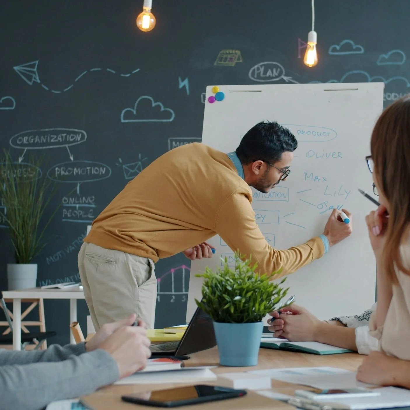 Man writing on whiteboard with colleagues sitting at a table in a modern office with chalk drawings on the wall.