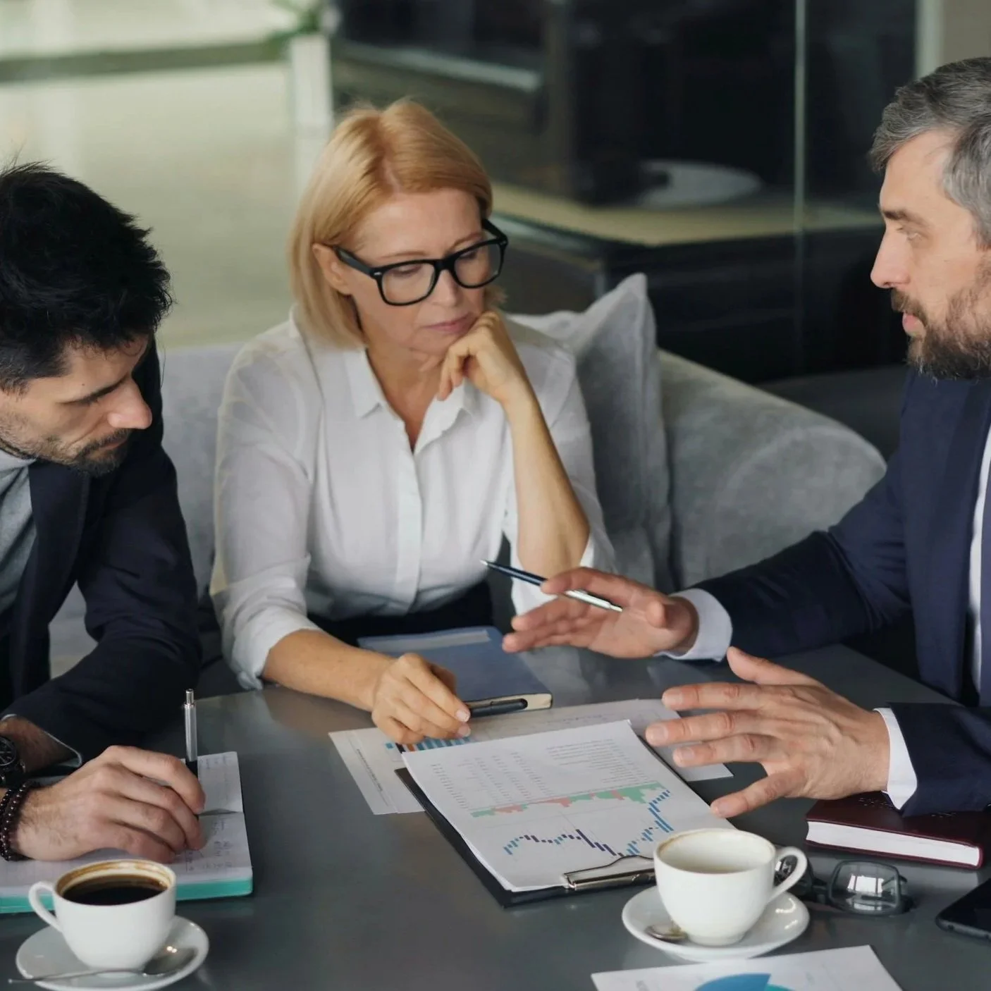 Three professionals, two men and one woman, engaged in a business discussion at a conference table with documents, charts, notebooks, pens, and coffee cups in a modern office setting.