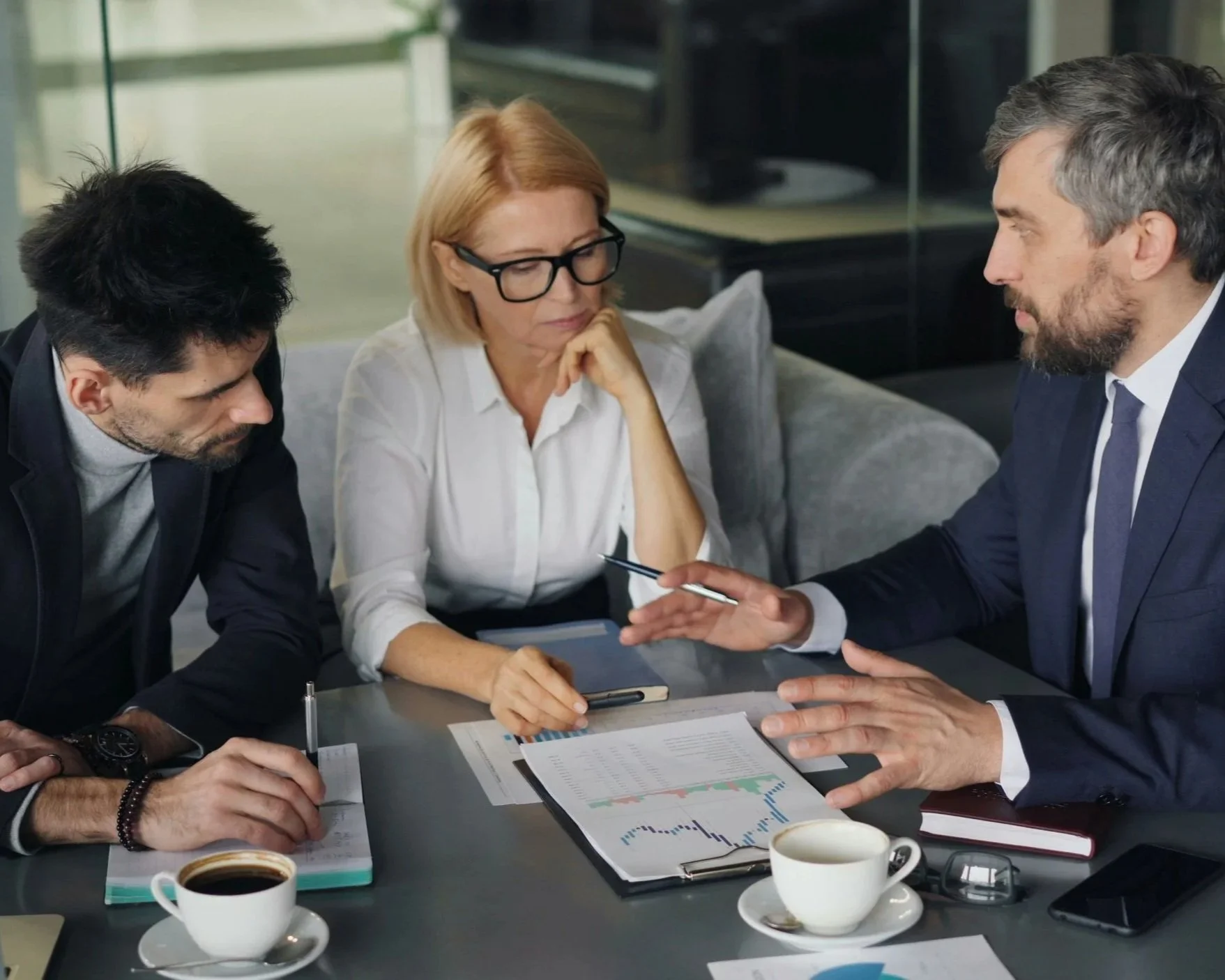 Three business professionals, two men and one woman, are engaged in a serious discussion at a conference table with coffee cups, documents, and electronic devices visible.