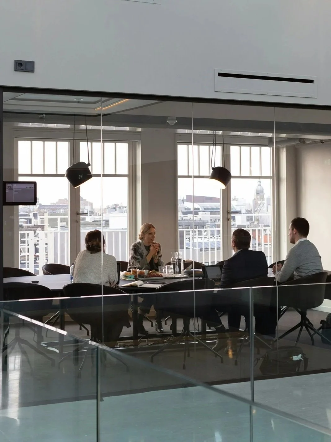 Four people sitting around a table in a modern office conference room, seen through glass walls, with city buildings visible outside the windows.