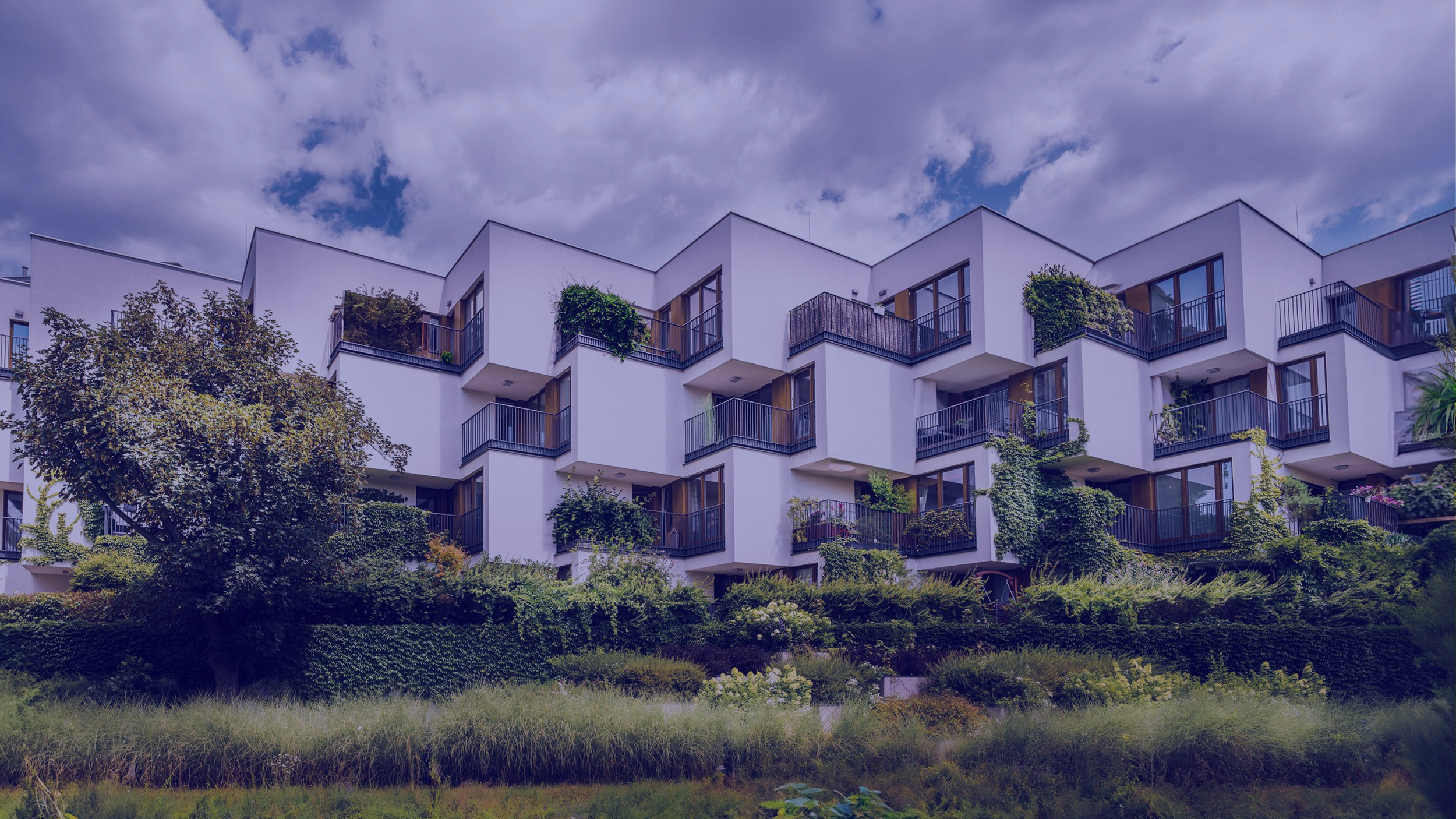 Modern white apartment building with multiple balconies, surrounded by trees and greenery, under a partly cloudy sky.