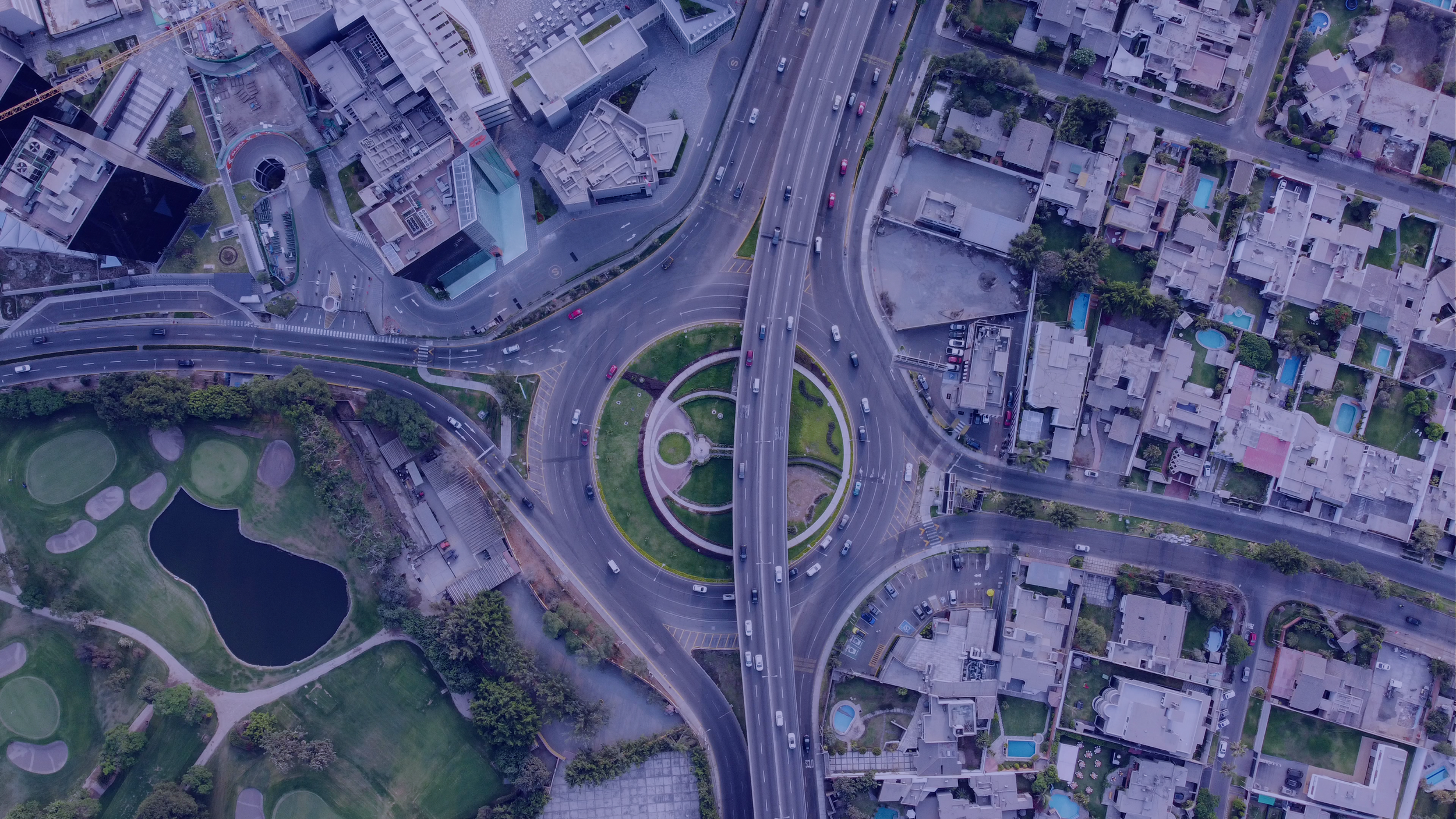 An aerial view of a city intersection with multiple roads, buildings, a park with a pond, and residential houses.